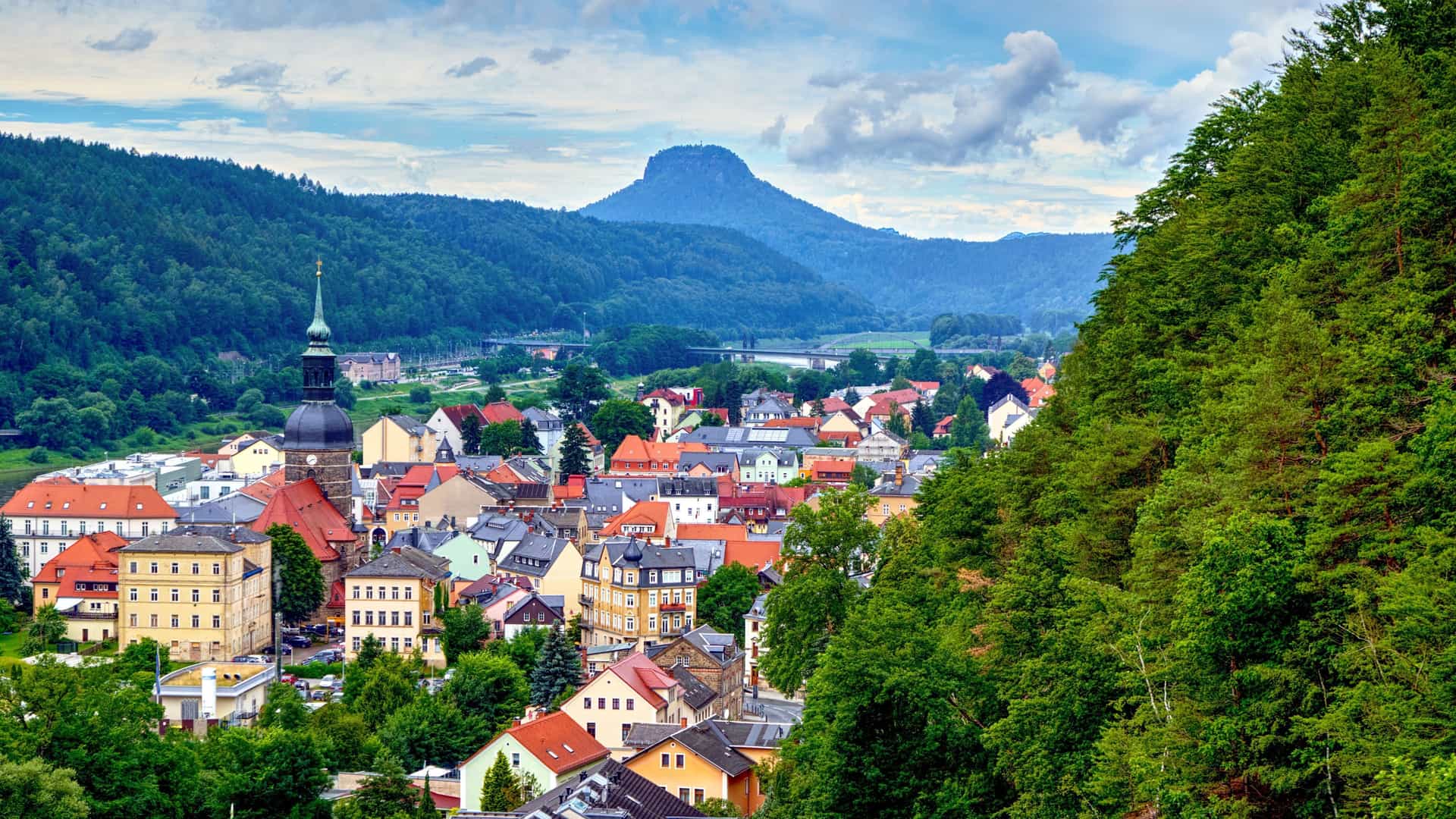 A picturesque view of the village of Bad Schandau in Germany, with colorful buildings and a church steeple in the foreground and a prominent mountain peak in the background.