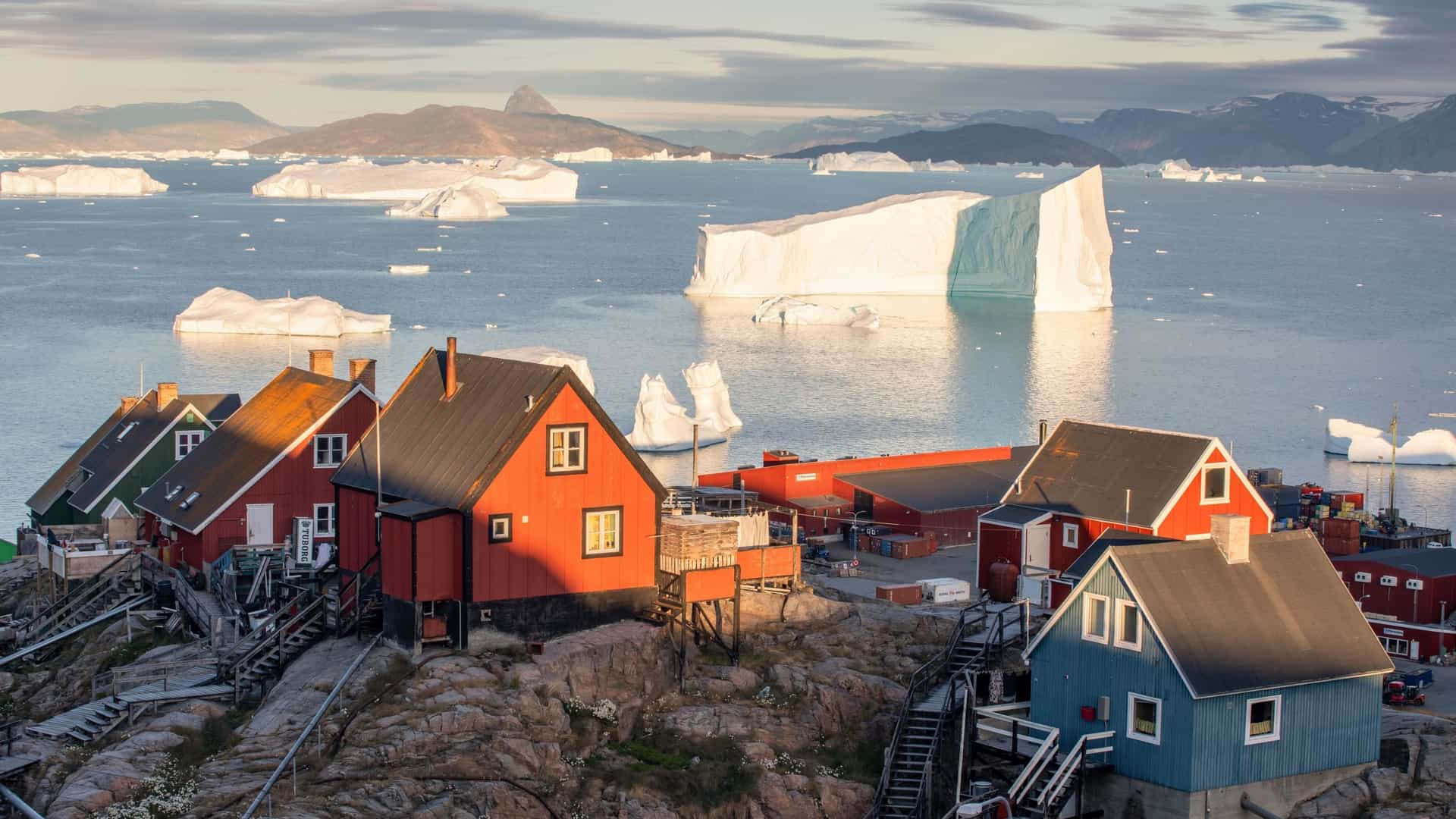 A remote Greenlandic village with colorful houses built on a rocky coastline, overlooking a fjord filled with massive icebergs in Baffin Bay.