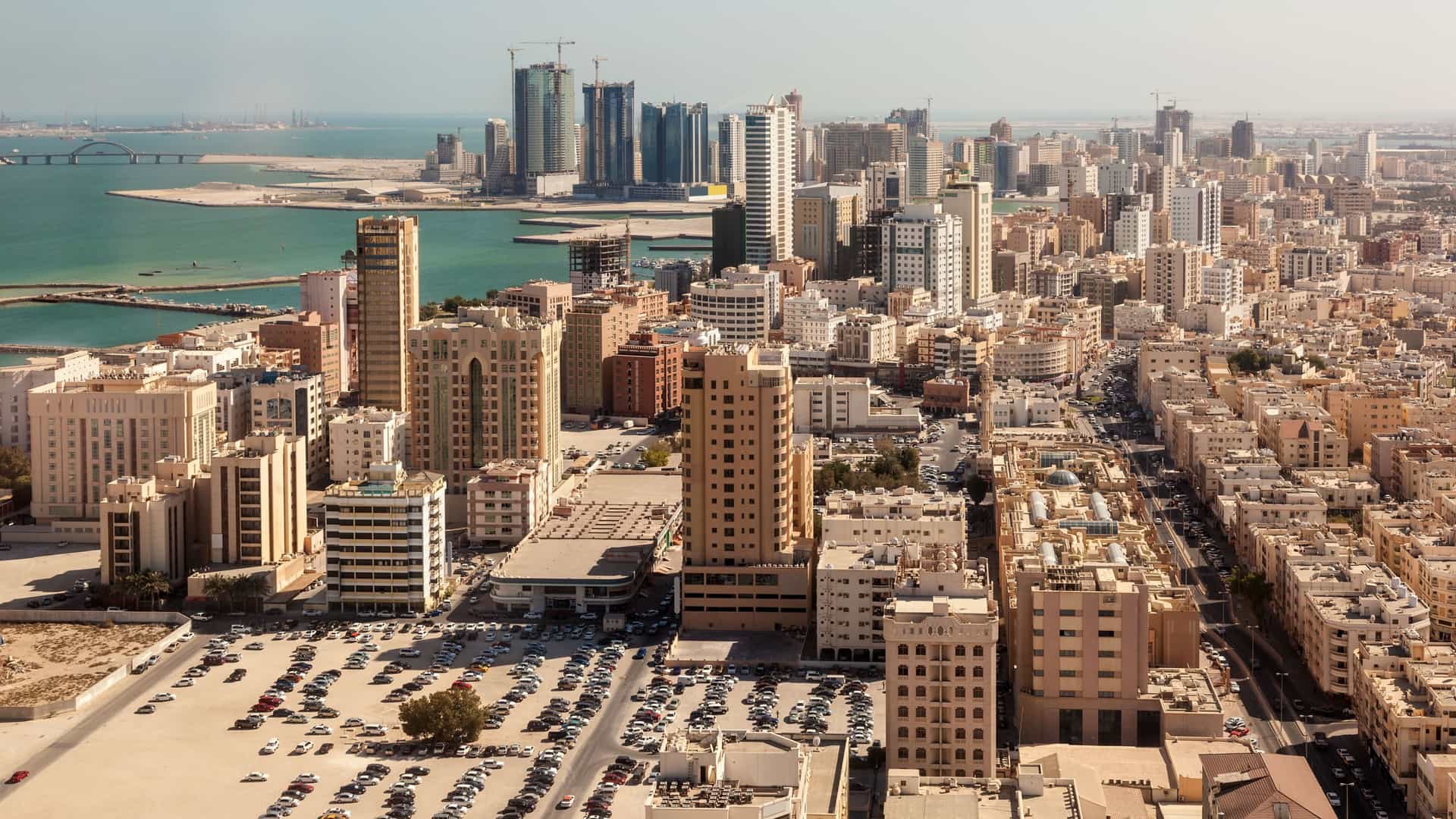 A high-angle aerial view of the modern cityscape of Manama, the capital of the Kingdom of Bahrain. The image shows a dense concentration of skyscrapers and high-rise buildings in the city's financial and residential districts, stretching along the coastline of the Arabian Gulf.