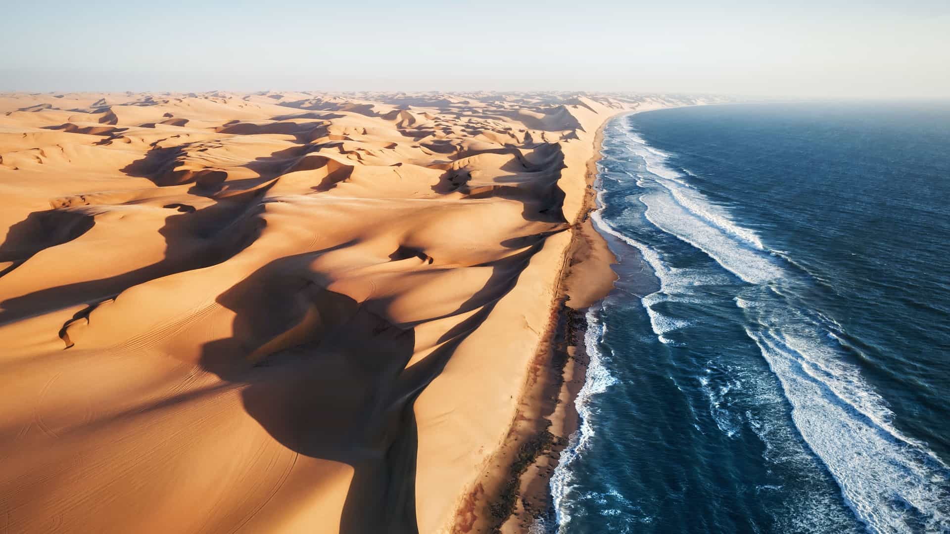 An aerial view of the dramatic, untouched landscape where the vast golden sand dunes of the Namib Desert meet the powerful blue waves of the Atlantic Ocean in Angola, Africa.
