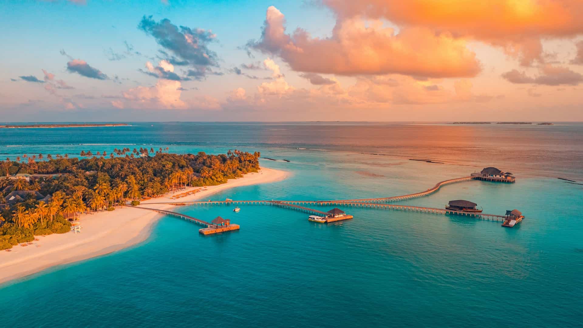 An aerial view of a tropical island resort at sunset, featuring white sand beaches, turquoise water, and long wooden piers leading to overwater bungalows.