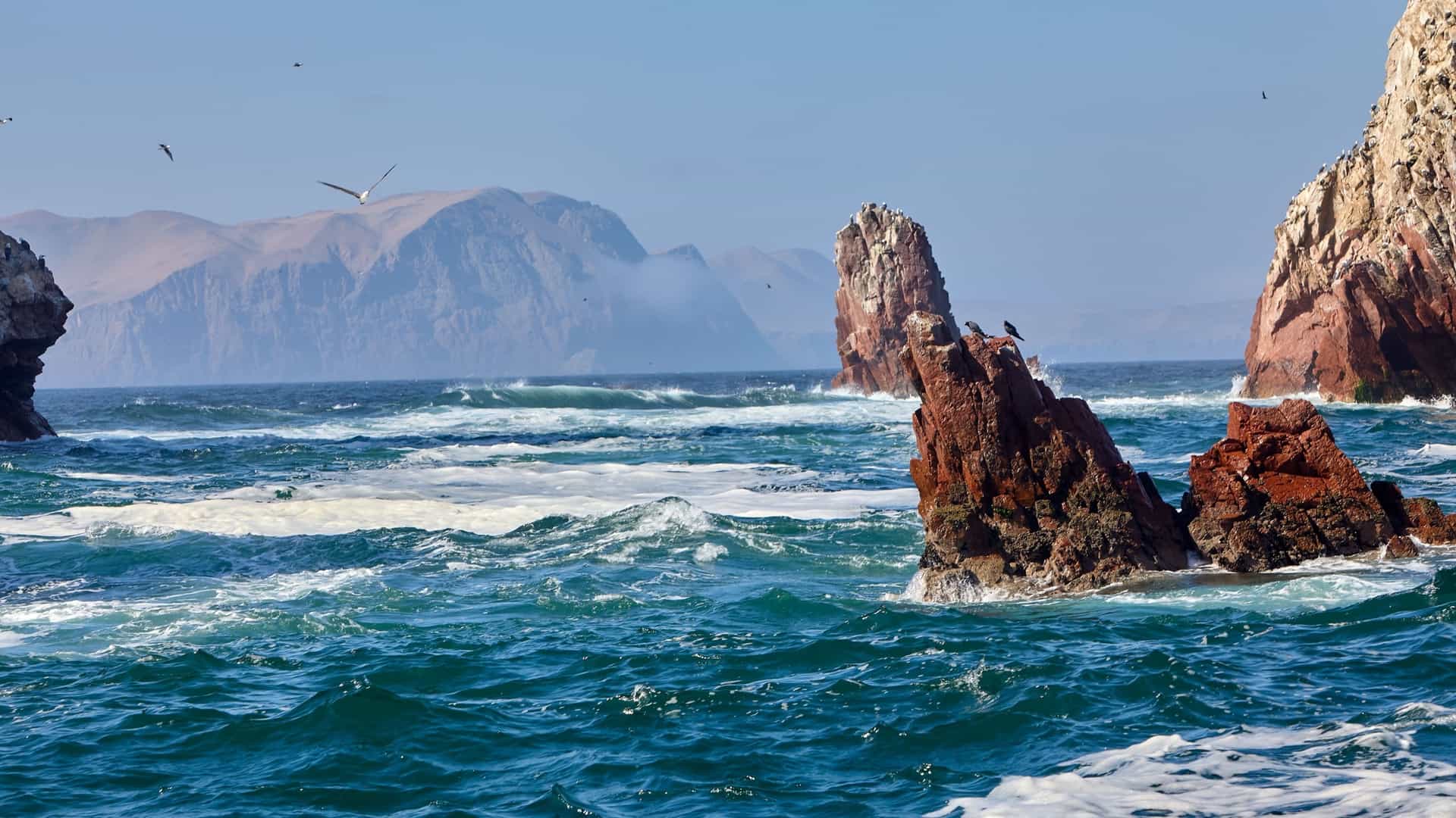 A scenic view of the Ballestas Islands in Peru, with waves crashing against jagged rock formations in the Pacific Ocean and a few birds flying overhead.