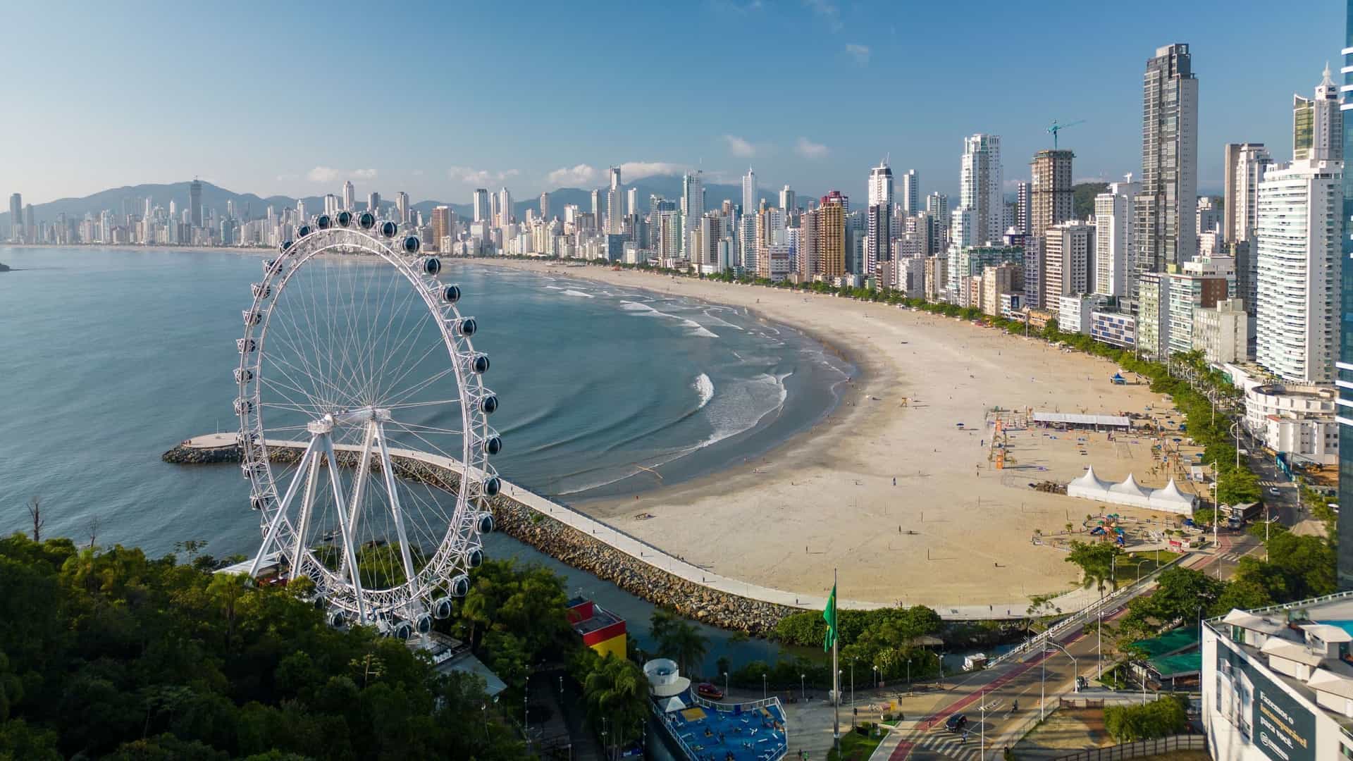 An aerial view of Balneário Camboriú's coastline, with a wide sandy beach, a large Ferris wheel on a pier, and a cityscape of towering high-rise buildings.