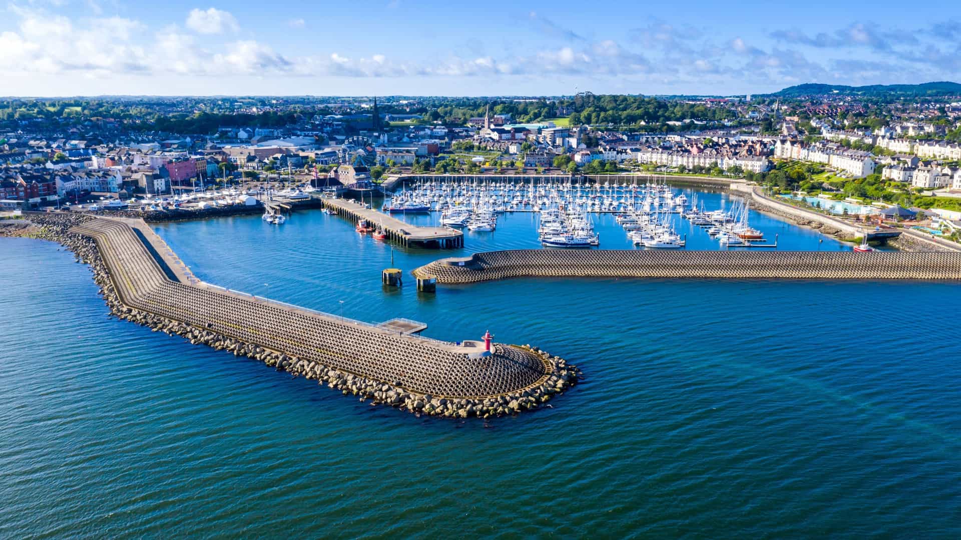 An aerial view of Bangor Marina in Northern Ireland, filled with boats, with a large breakwater protecting the harbor and a cityscape in the background.