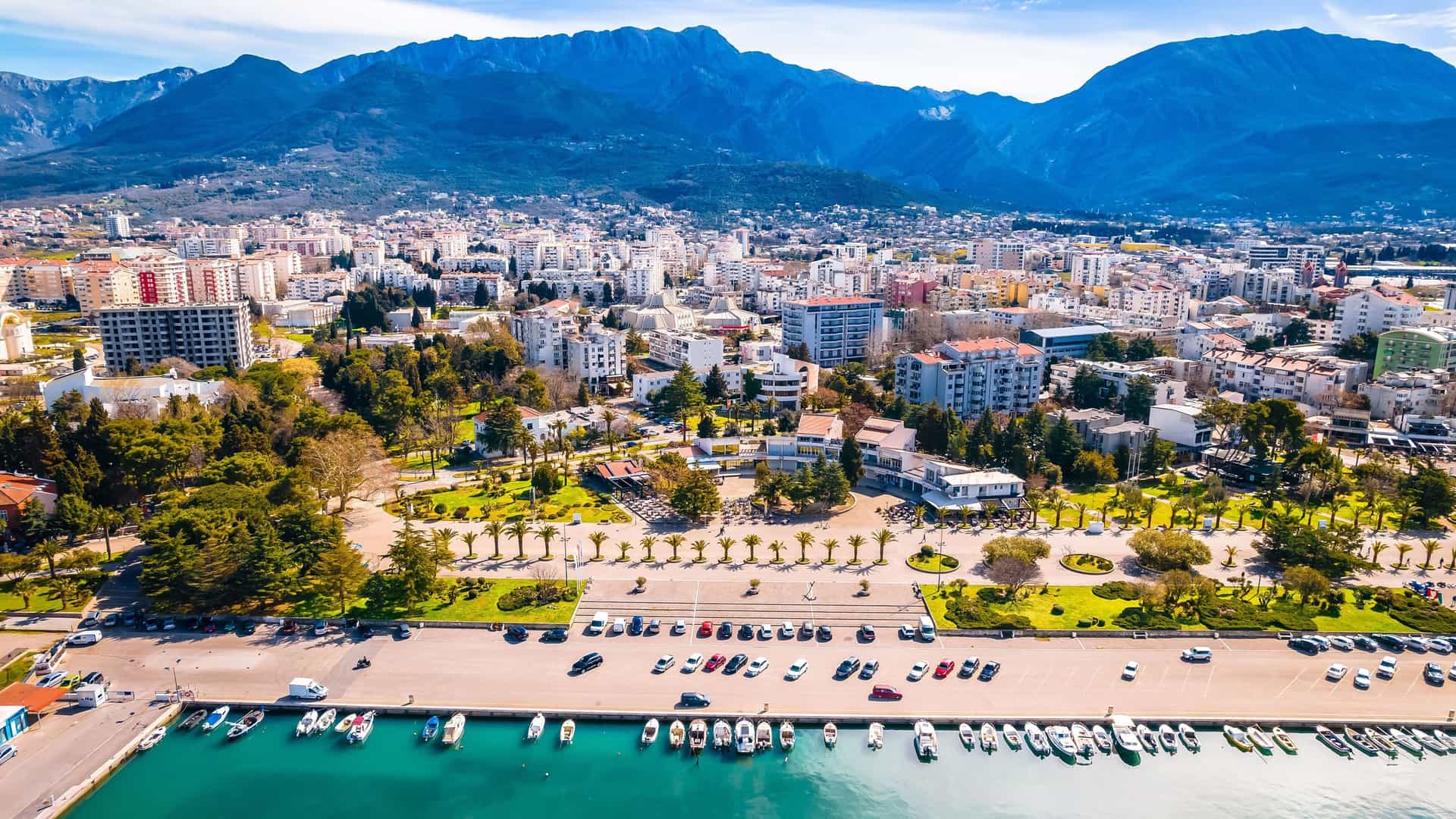 An aerial view of Bar, Montenegro, showing the bustling marina filled with boats, a waterfront promenade, and the modern city against a backdrop of majestic mountains.