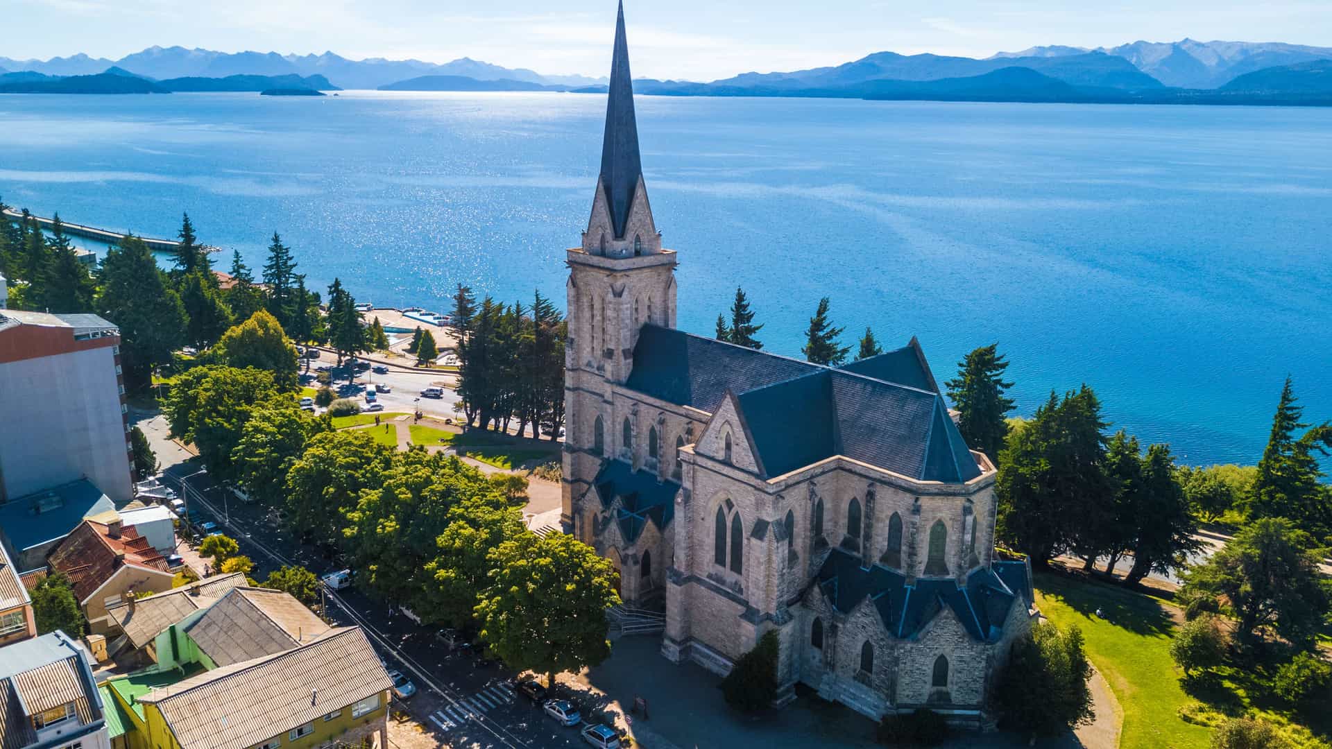 An aerial view of Bariloche, Argentina, showing the prominent stone Catedral de San Carlos de Bariloche with its tall spire. The church is situated on the shore of the deep blue Nahuel Huapi Lake, which stretches toward a distant range of hazy mountains.