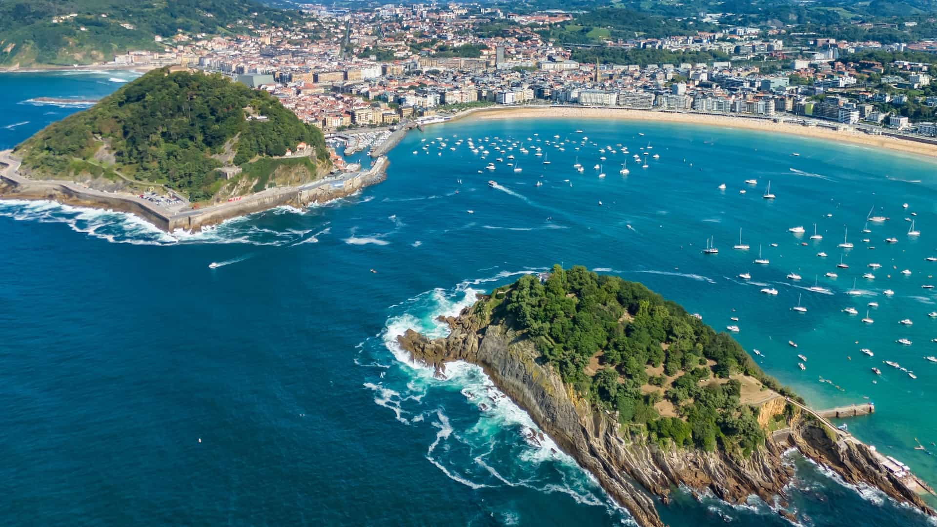 An aerial view shows the bay of San Sebastián in the Basque Region of Spain, featuring boats and two lush green islands.