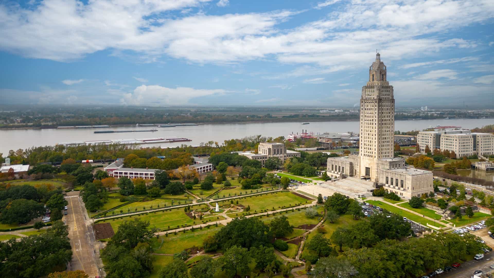 An aerial view of the iconic Louisiana State Capitol building and surrounding gardens, with the Mississippi River and industrial barges visible in the background.