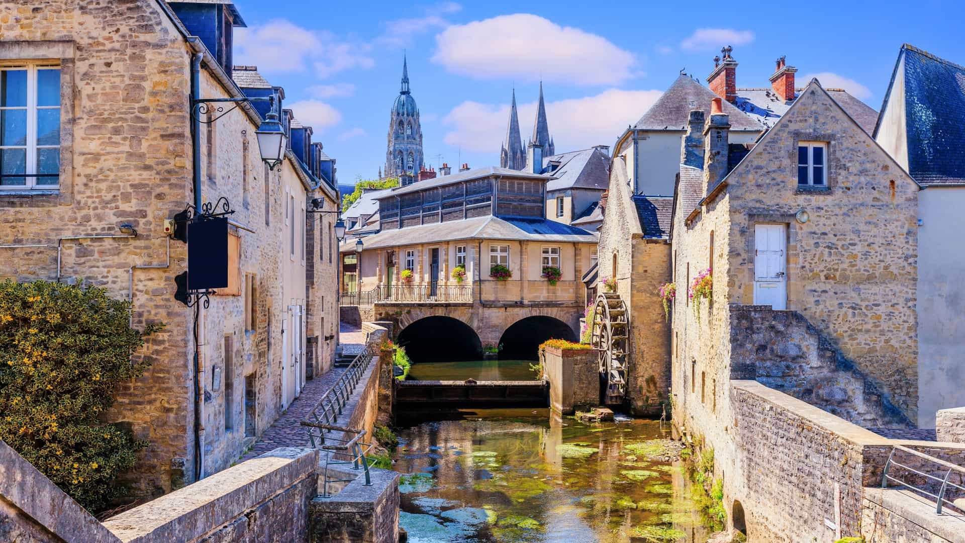 A scenic canal flows through the historic streets of Bayeux, lined with picturesque stone buildings and a towering cathedral in the distance.