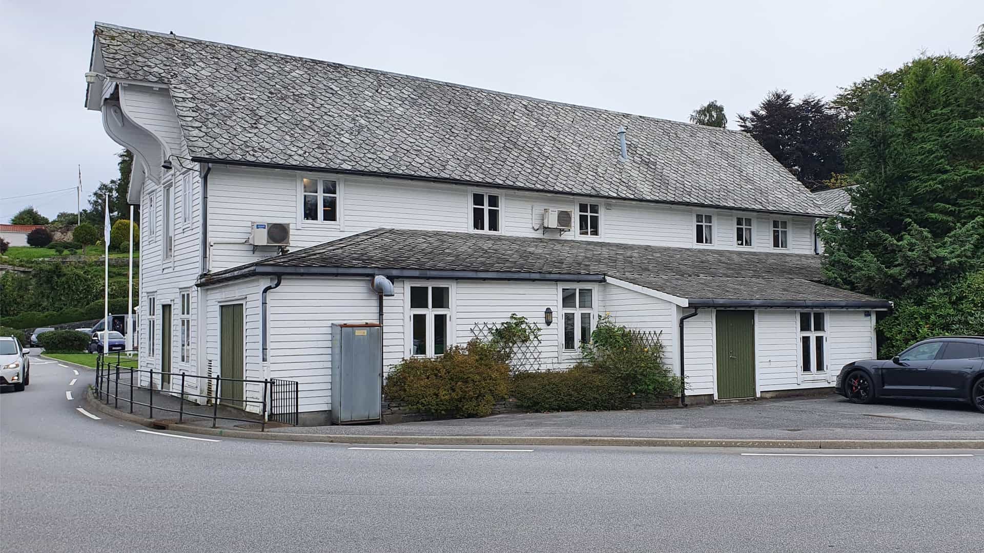 A traditional white wooden building with a slate roof in the small Norwegian village of Bekkjarvik, nestled by the coast with lush greenery and a paved road in front.