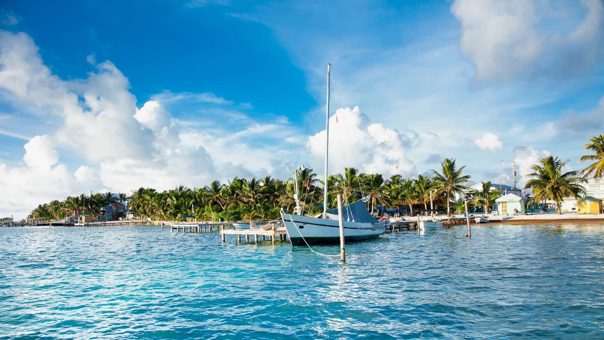 A beautiful sailboat rests in the shallow, clear turquoise waters off the coast of Belize City, with lush palm trees and colorful buildings lining the shore.