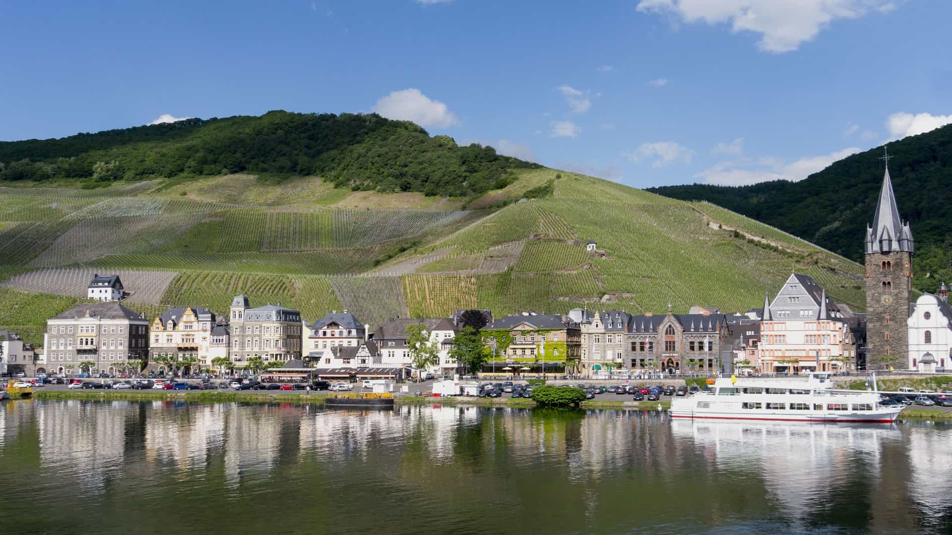 A beautiful landscape of Bernkastel-Kues, Germany, with a line of buildings and a ferry along the Moselle River. Terraced vineyards climb the steep hillside behind the town.