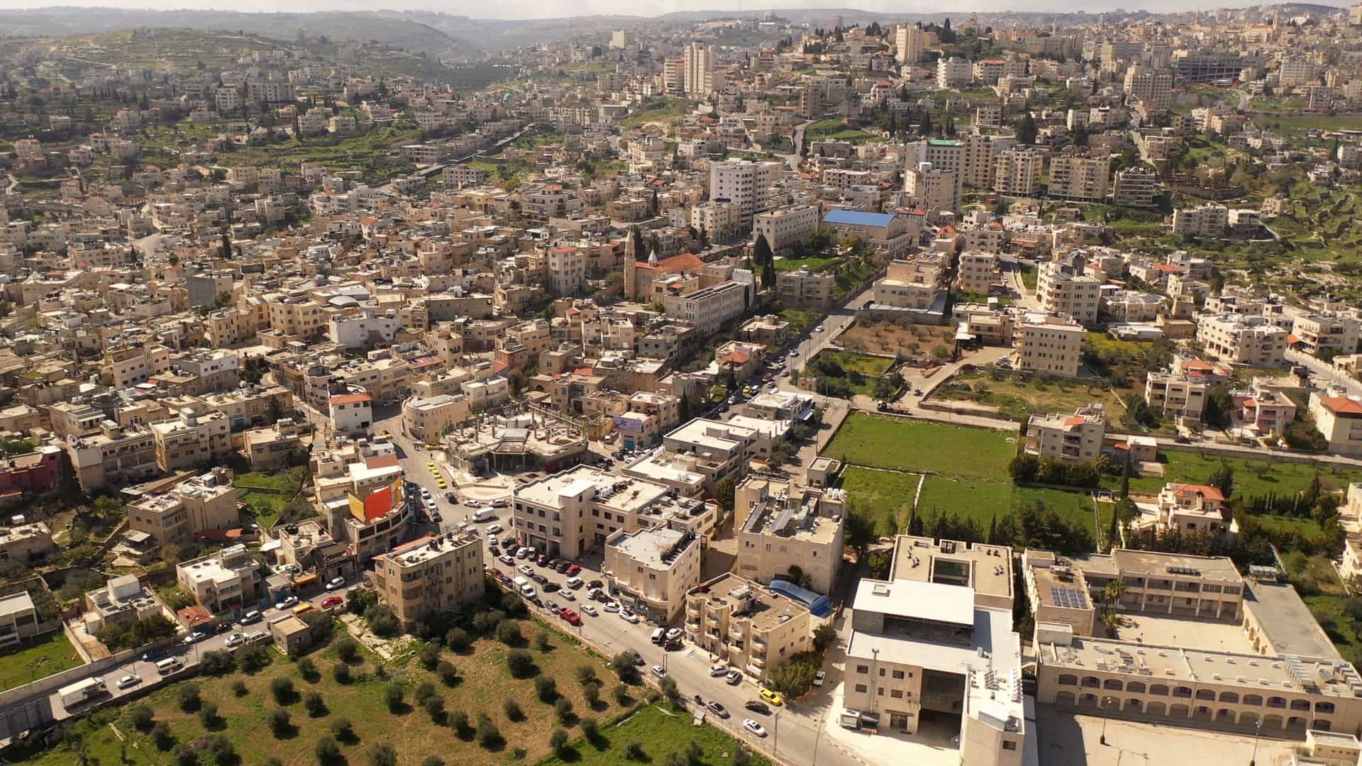 An aerial, drone view of Bethlehem City in the Palestinian Authority, captured in the morning. The image shows a dense urban landscape of buildings interspersed with green fields and hills in the background. The city's structures are a mix of traditional and modern construction.