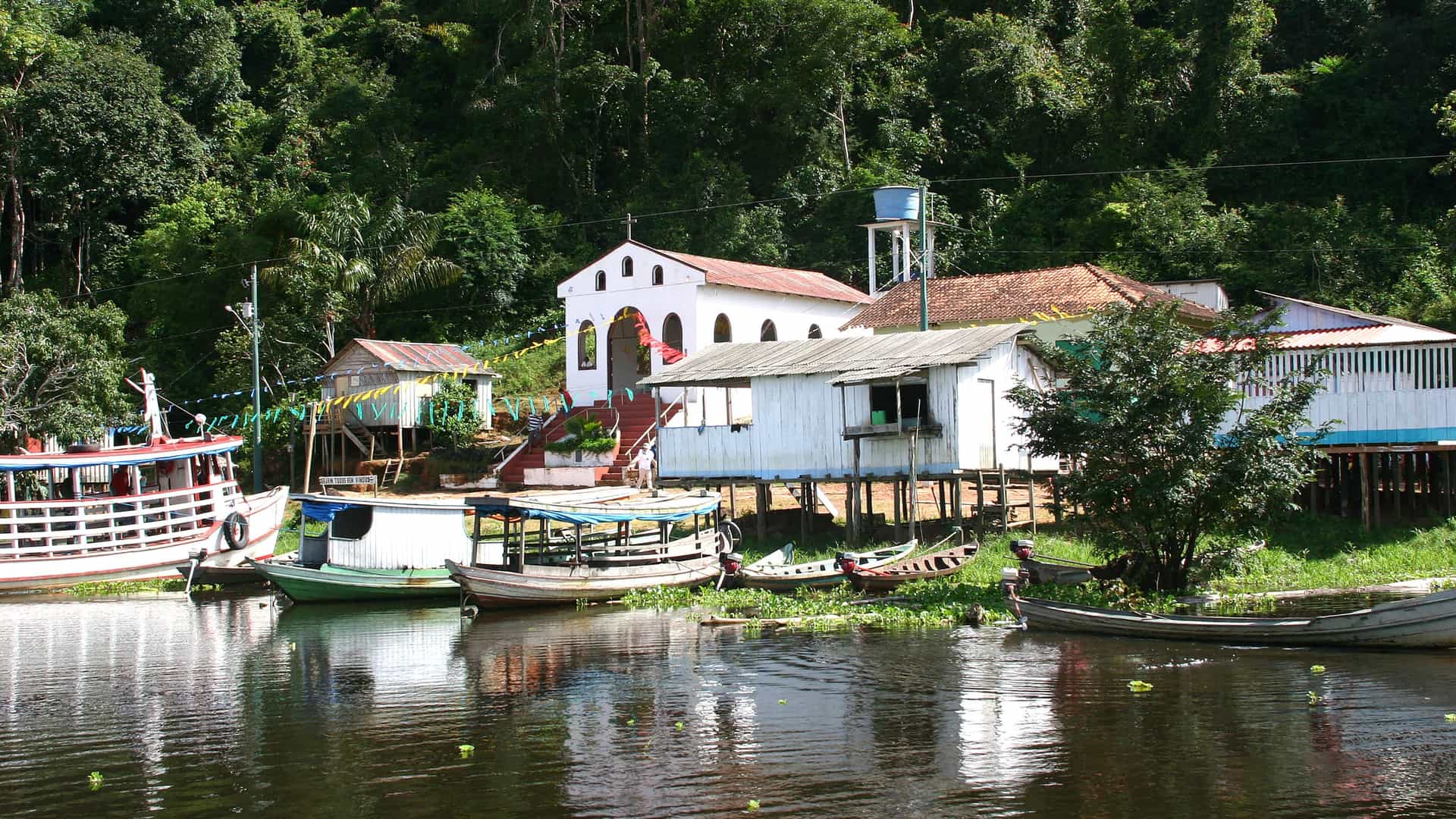 A scenic view of Boca da Valeria, a small riverside village on the Amazon River, with several boats docked in front of colorful houses and lush rainforest.