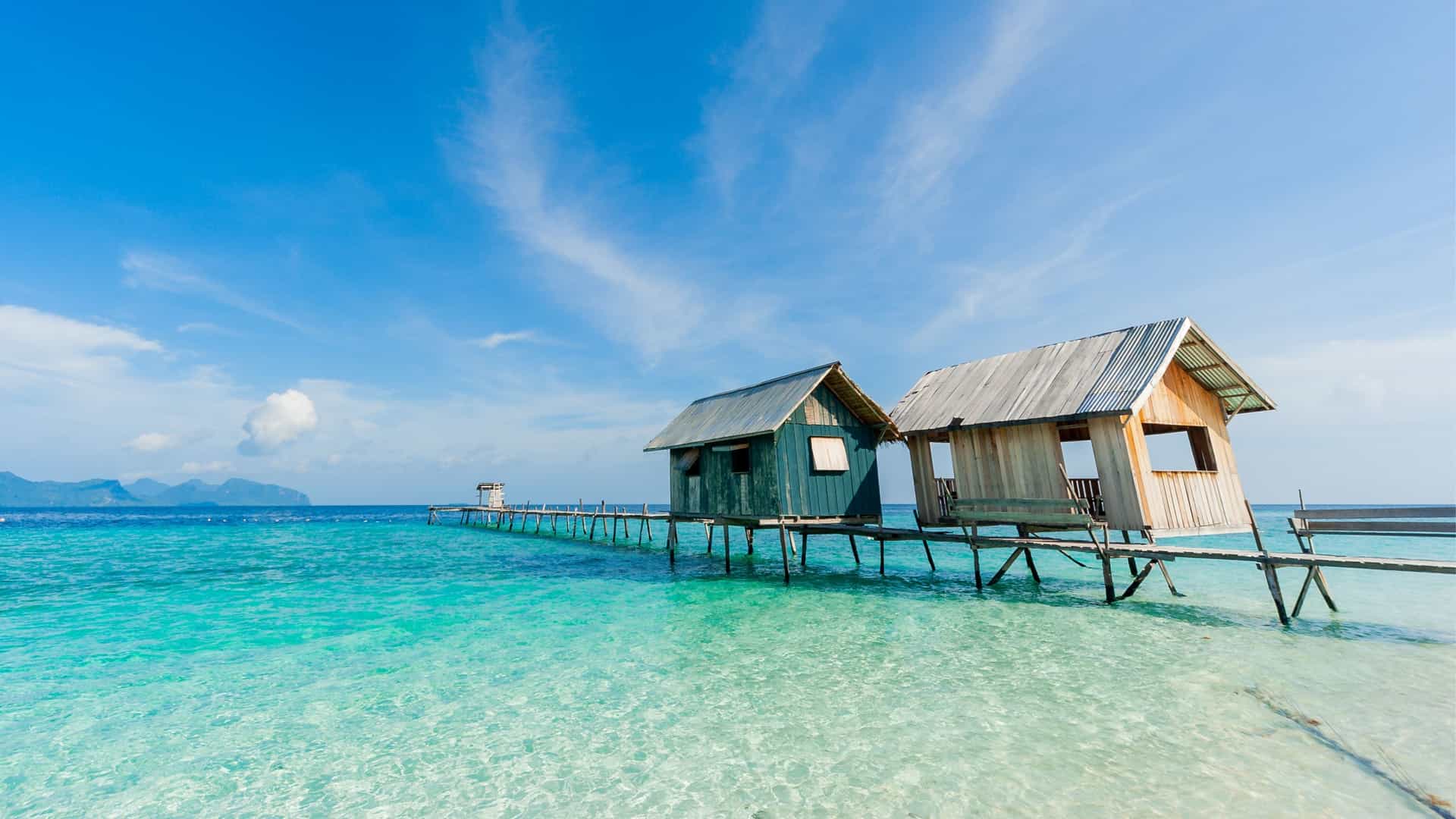 Two wooden stilt houses sit on a pier over the clear turquoise water of Bohey Dulang Island, Malaysia.