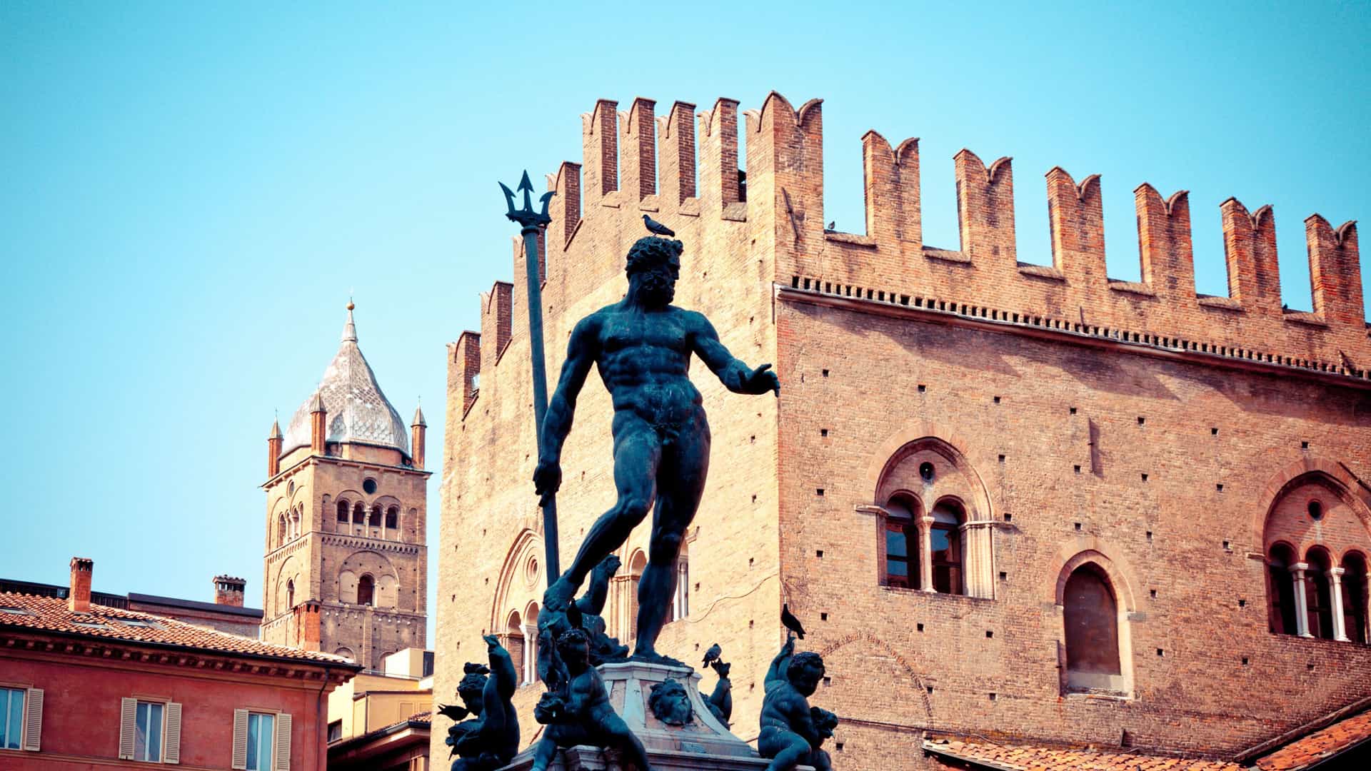 A detailed shot of the historic bronze Fountain of Neptune statue in Bologna, Italy, with the medieval Palazzo del Podestà and its iconic clock tower visible in the background.