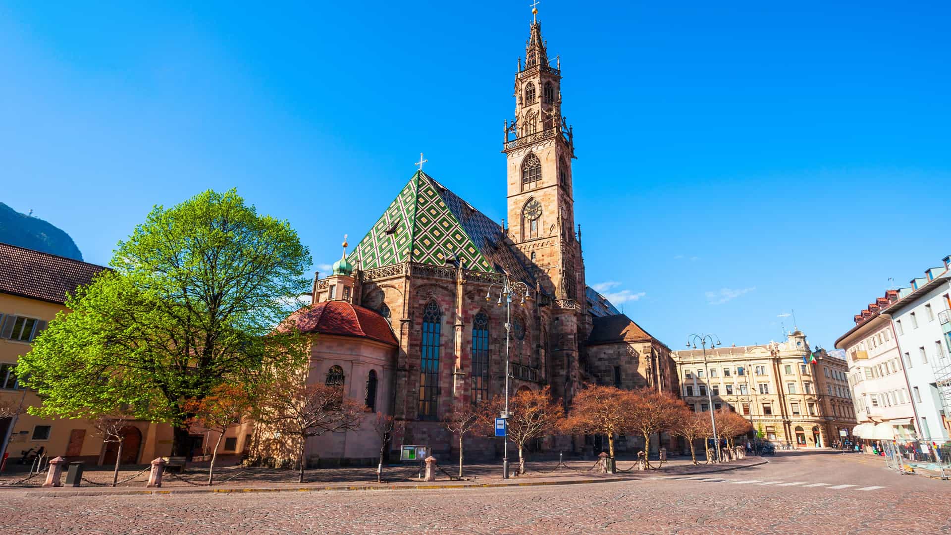 The Bolzano Cathedral in Bolzano, Italy, stands prominently on a wide, cobblestone square under a vibrant blue sky. The Gothic-style cathedral features a tall, intricately carved spire and a distinctive roof with a green and yellow diamond pattern. In the foreground, a tree with bright spring leaves and other bare trees line the square.