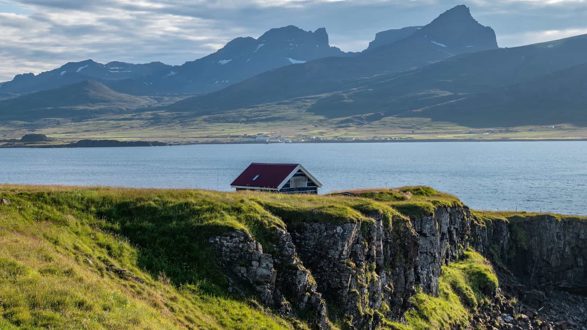 A scenic, wide-angle view of the small fishing village of Bakkagerði, located in the Borgarfjörður Eystri fjord in East Iceland. The village features colorful houses clustered along the coastline next to a calm bay of the sea, which is surrounded by steep, rugged, snow-dusted mountains under a clear sky.