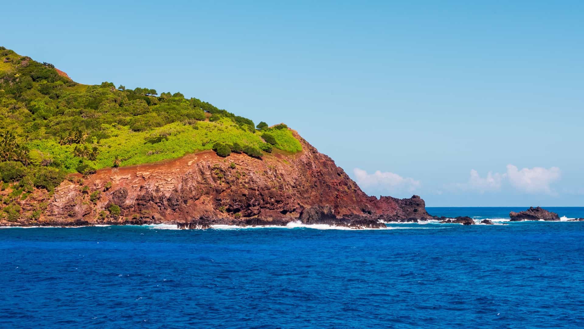 A photograph of Bounty Bay on Pitcairn Island, a British Overseas Territory in the South Pacific. The small bay is famous for being the final resting place of the HMS Bounty, which was burned by the mutineers who settled the island. The image shows the steep, rocky coastline of the island plunging down to the turquoise ocean, with a small landing area or dock visible at the edge of the bay. The island's slopes are covered in lush green tropical vegetation.