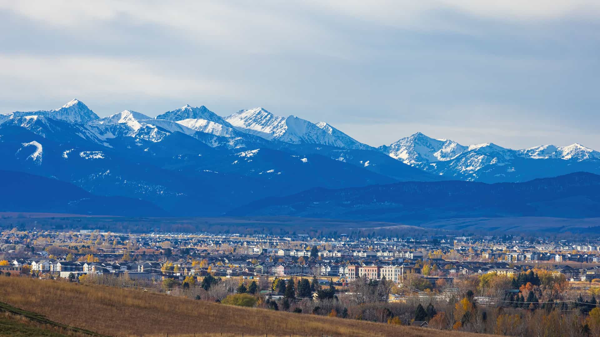 A panoramic view of Bozeman, Montana, with the snow-capped peaks of the Bridger Mountains dominating the background. The town fills the midground, with houses and buildings surrounded by trees displaying yellow and orange autumn foliage. A brown, grassy hill is in the foreground under a partially cloudy blue sky.