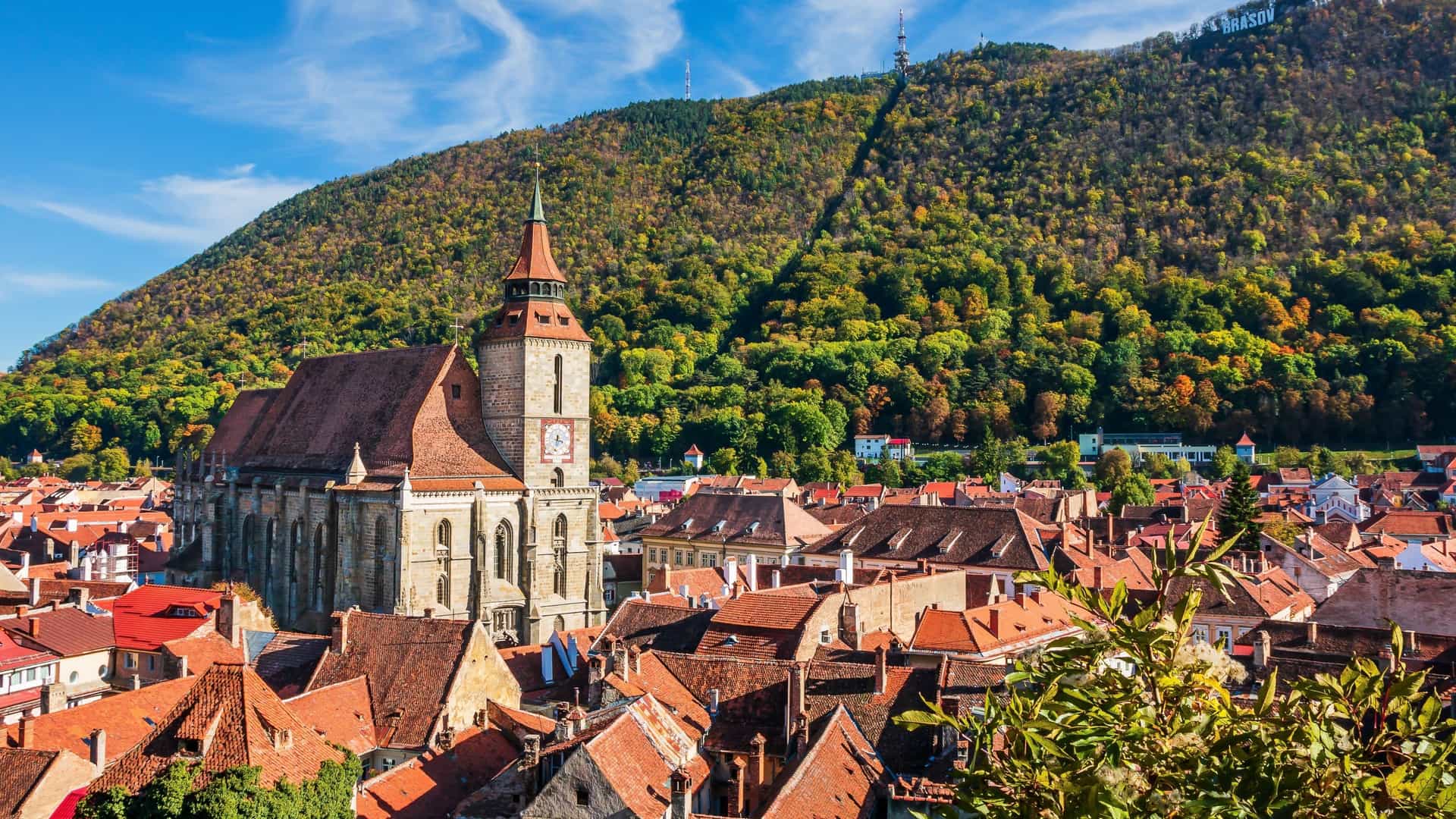 A beautiful elevated view of the historic Black Church (Biserica Neagră) and the red-tiled roofs of the old town in Brașov, Romania, nestled against a tree-covered mountain with the city name spelled out on its side.