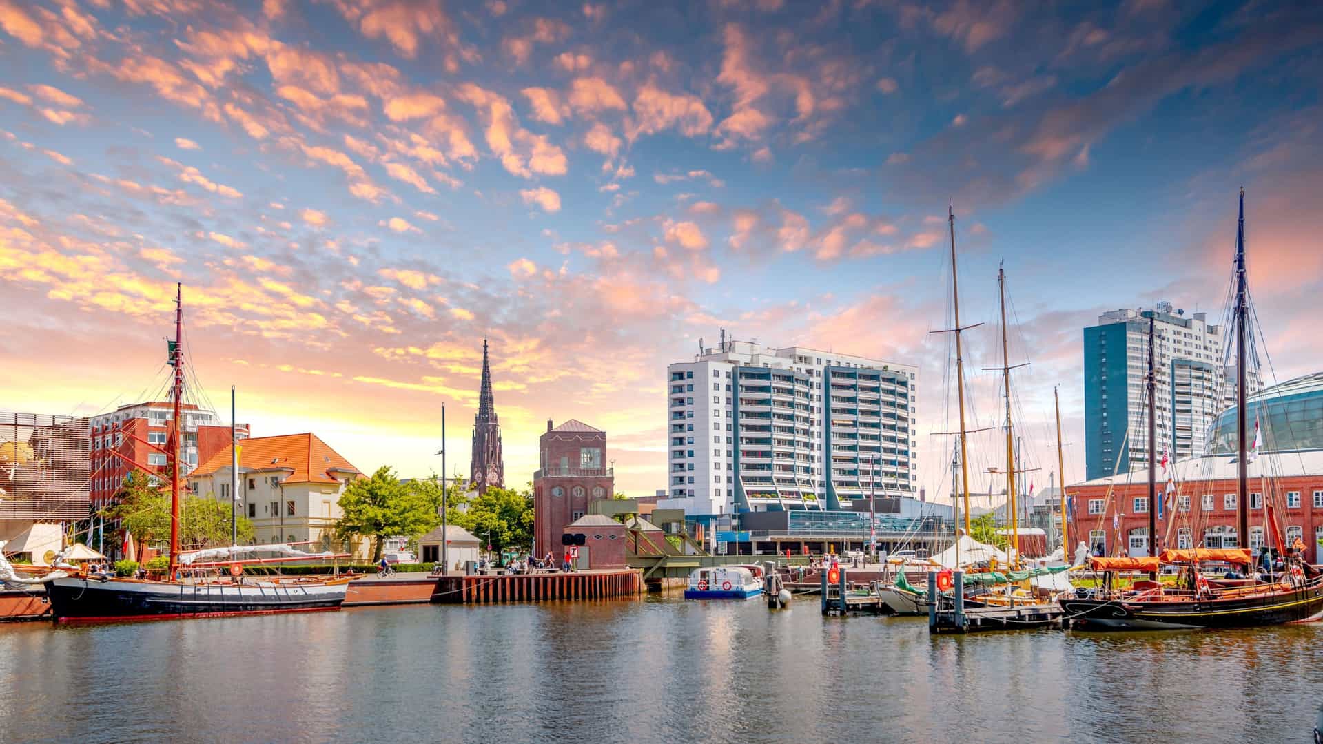 A picturesque sunset view of the harbor in Bremerhaven, Germany, featuring historic sailing ships docked along the waterfront with modern buildings and a church steeple silhouetted against the vibrant sky.