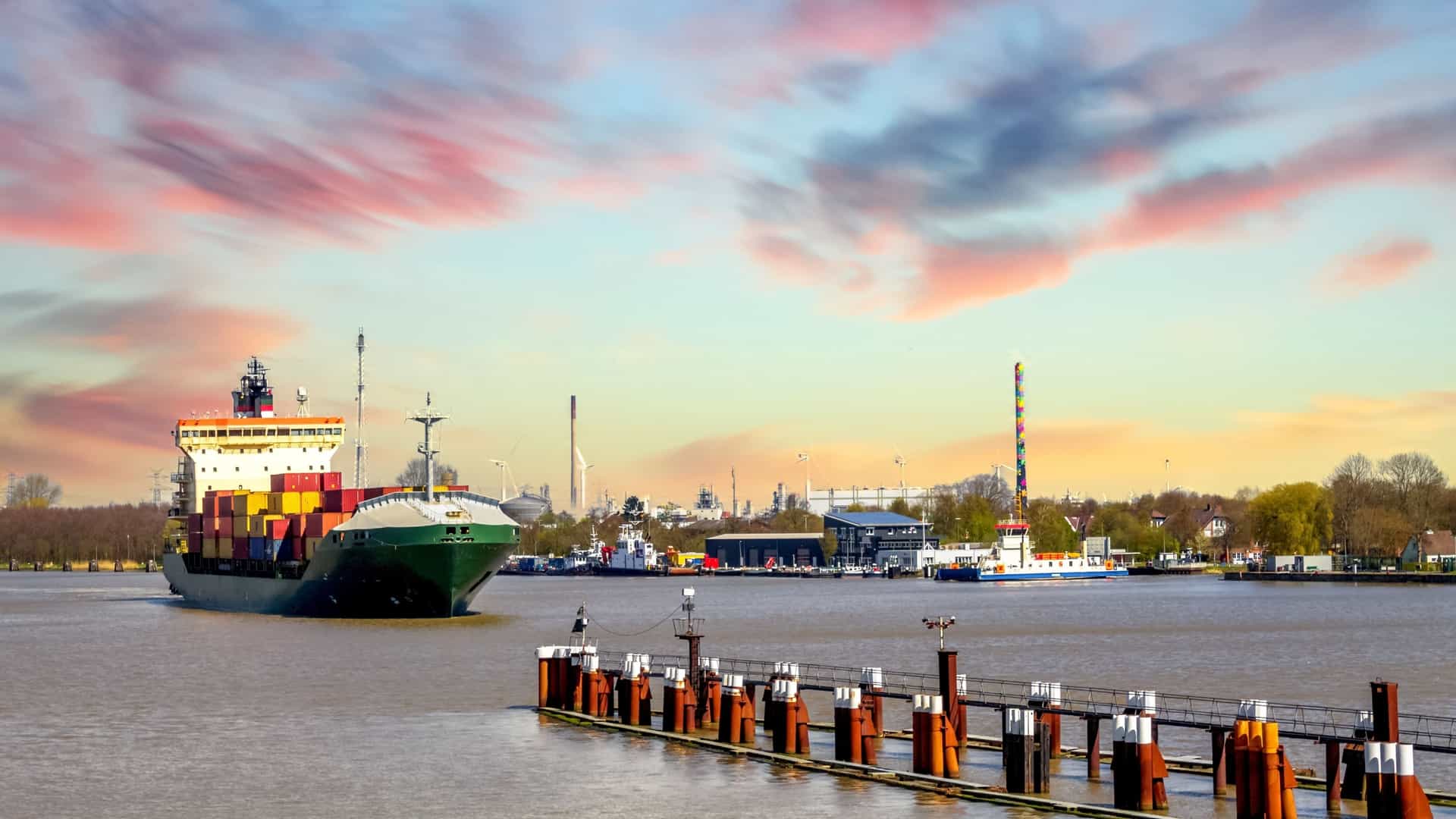 A large, green container ship moves along the water near a wooden dock in the harbor of Brunsbüttel, Germany, under a colorful sunset sky.