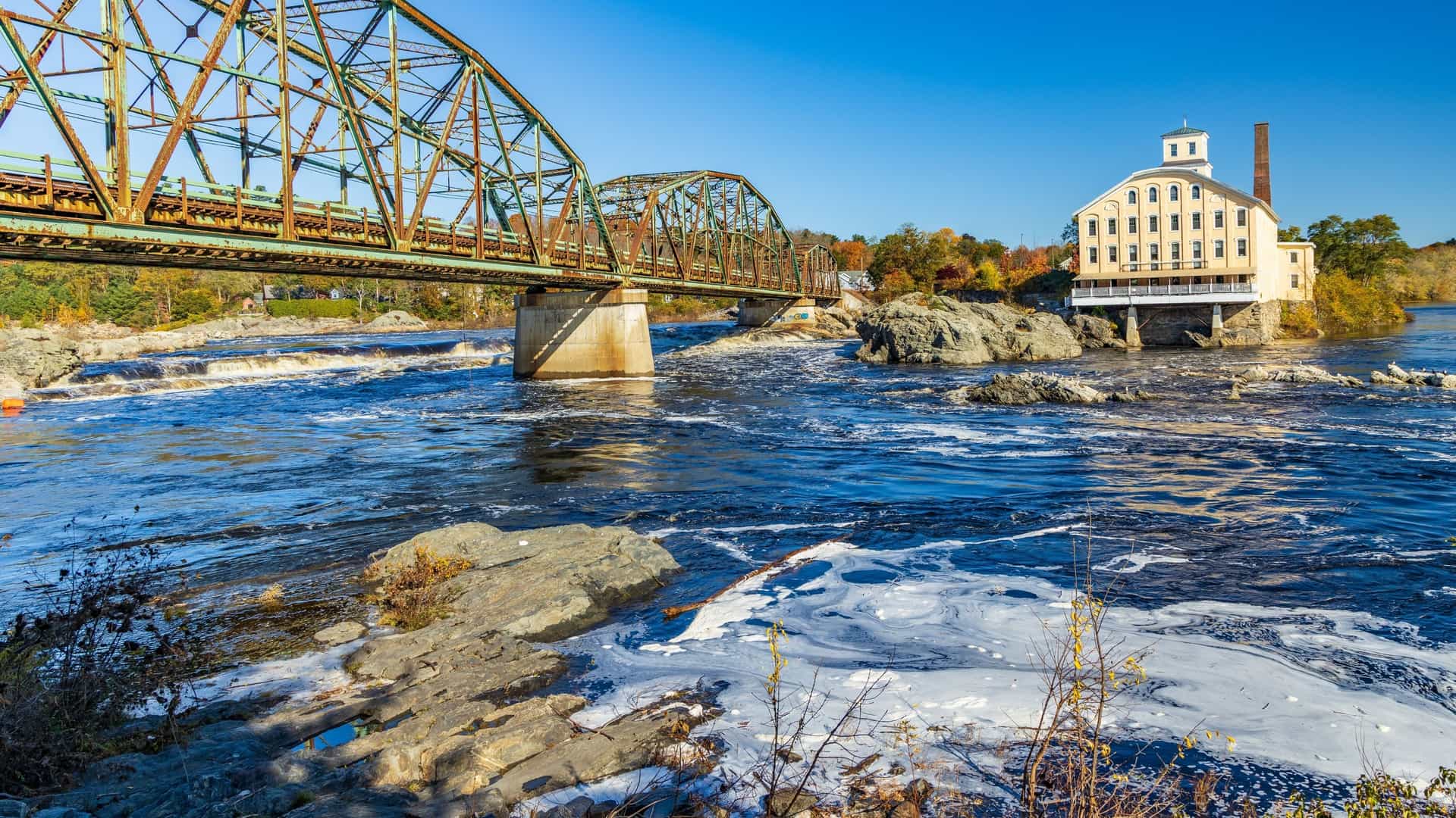A view of a truss bridge arching over the Androscoggin River with a historic mill building on the far bank in Brunswick, Maine.