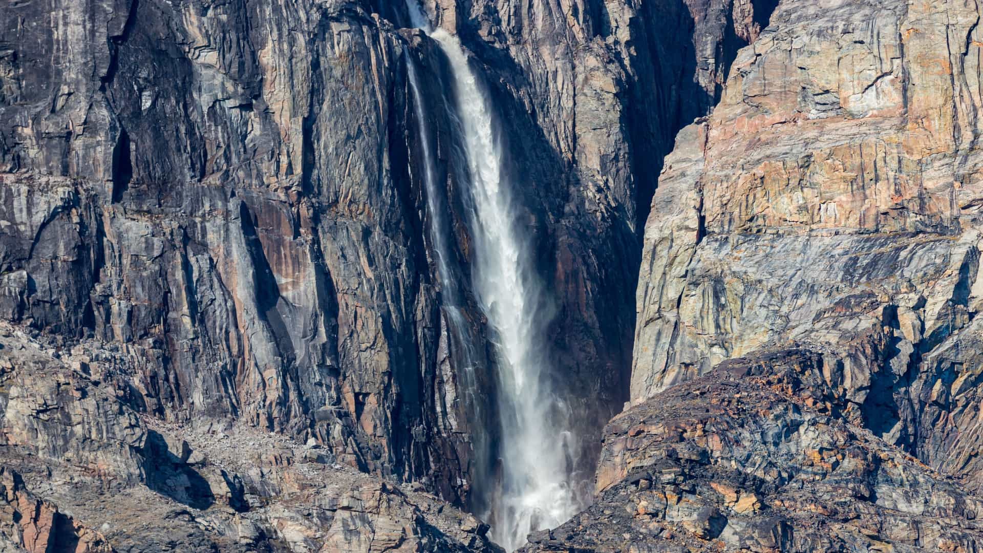 A large, powerful waterfall cascades down a sheer, rocky cliff face in Buchan Gulf, Nunavut, Canada.