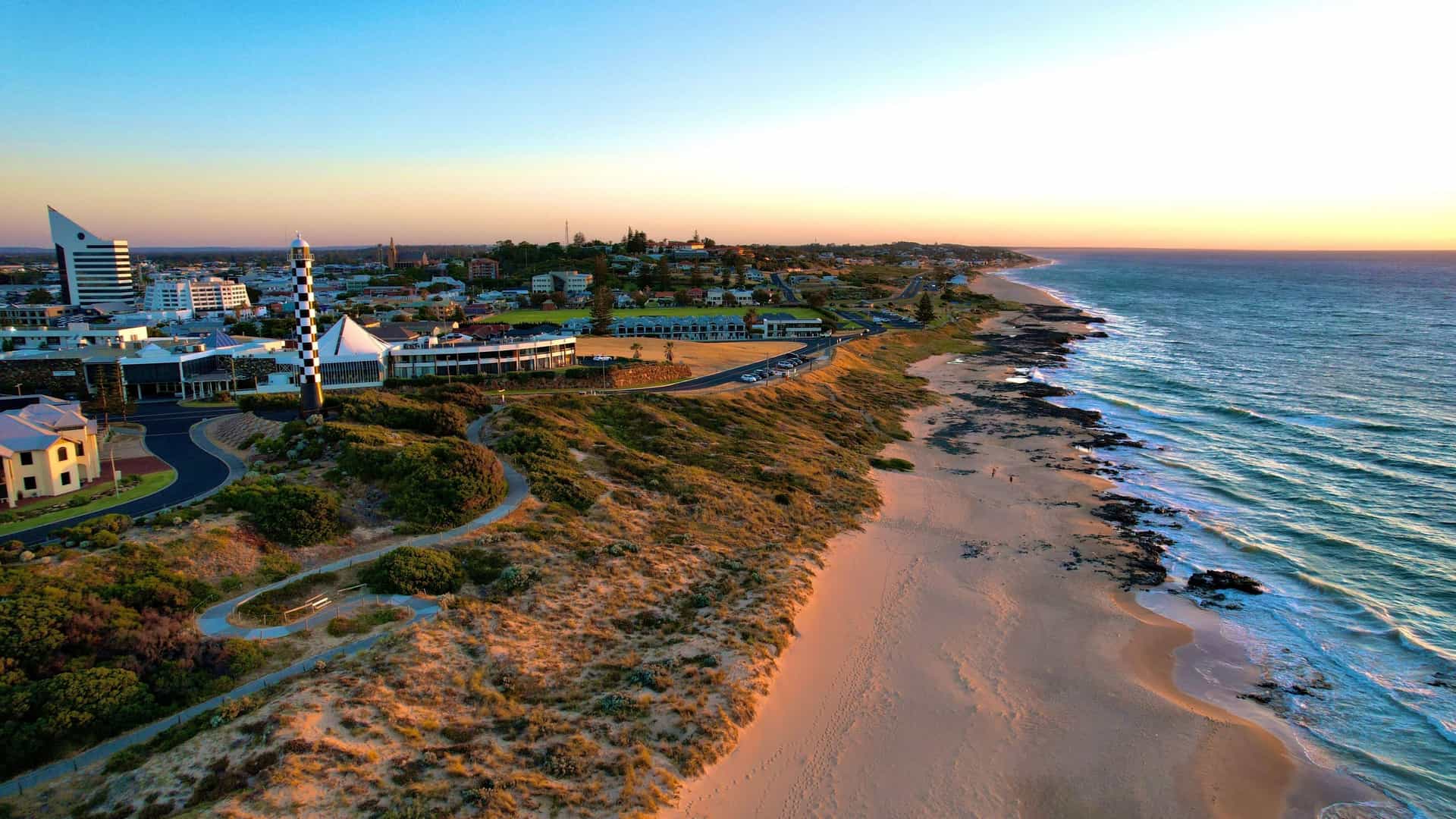 An aerial view of Bunbury, Western Australia, at sunset, showing the coast, city skyline, and the prominent striped lighthouse.