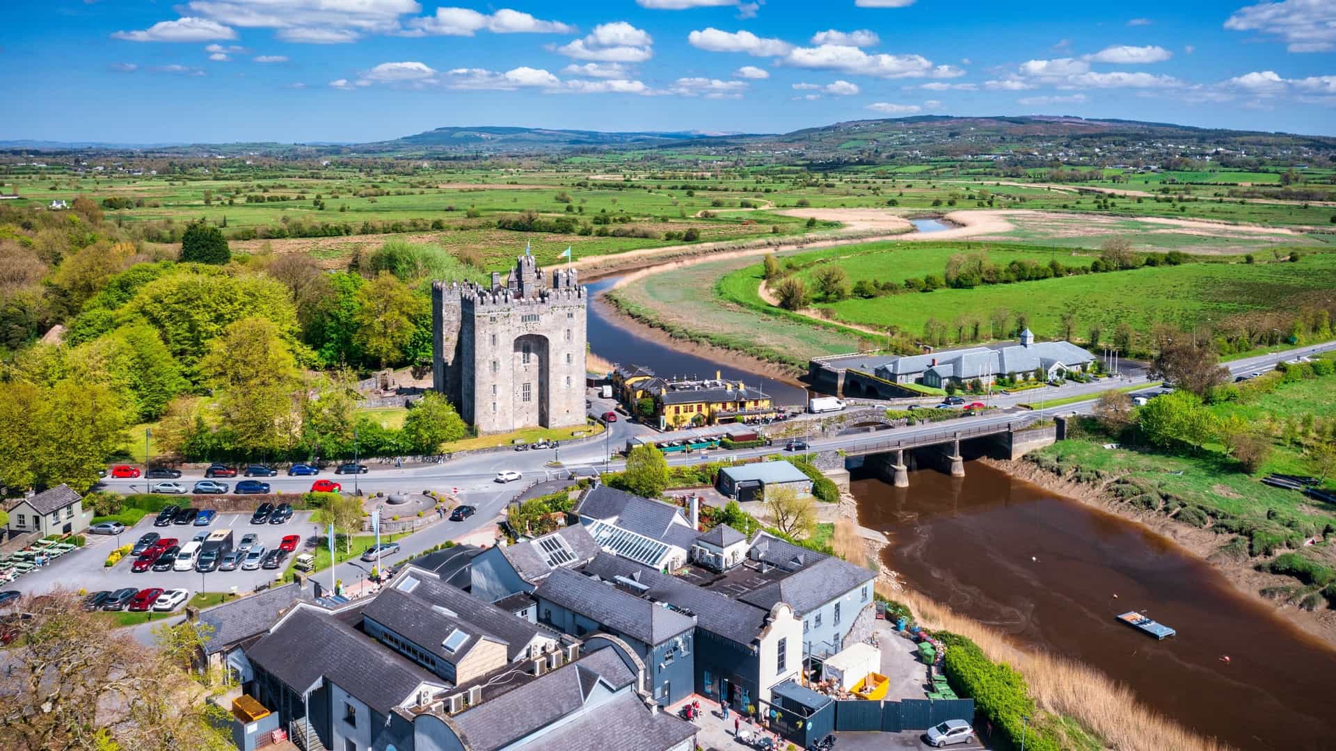 A dramatic, wide-angle view of the 15th-century Bunratty Castle in County Clare, Ireland. The large, imposing stone tower house dominates the scene, with its tall rectangular structure and corner turrets. It is set against a vibrant green lawn and a backdrop of trees, with a bright blue sky overhead featuring scattered white clouds. The castle's grey stone is well-preserved, reflecting its status as one of Ireland's most complete medieval fortresses.