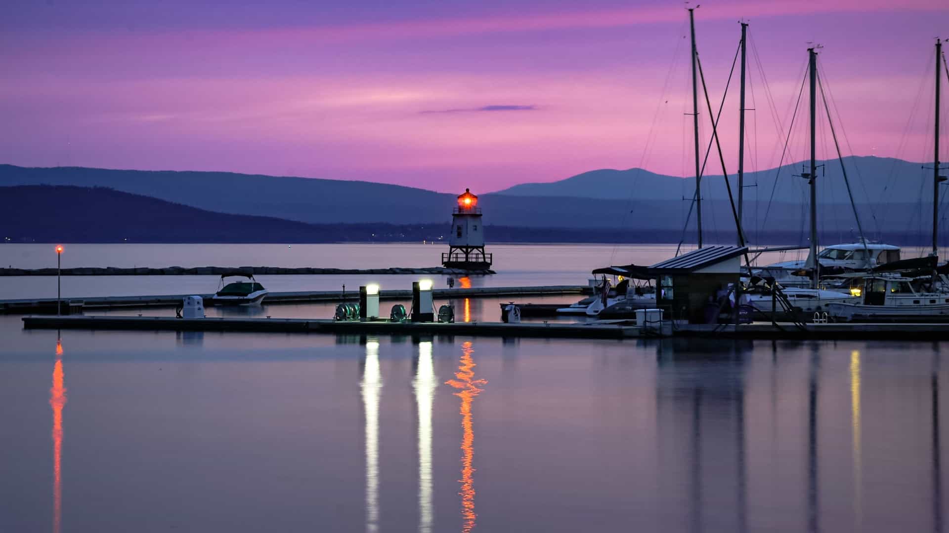 A tranquil dusk view of the Burlington harbor on Lake Champlain, with a lighthouse and sailboats at the dock.