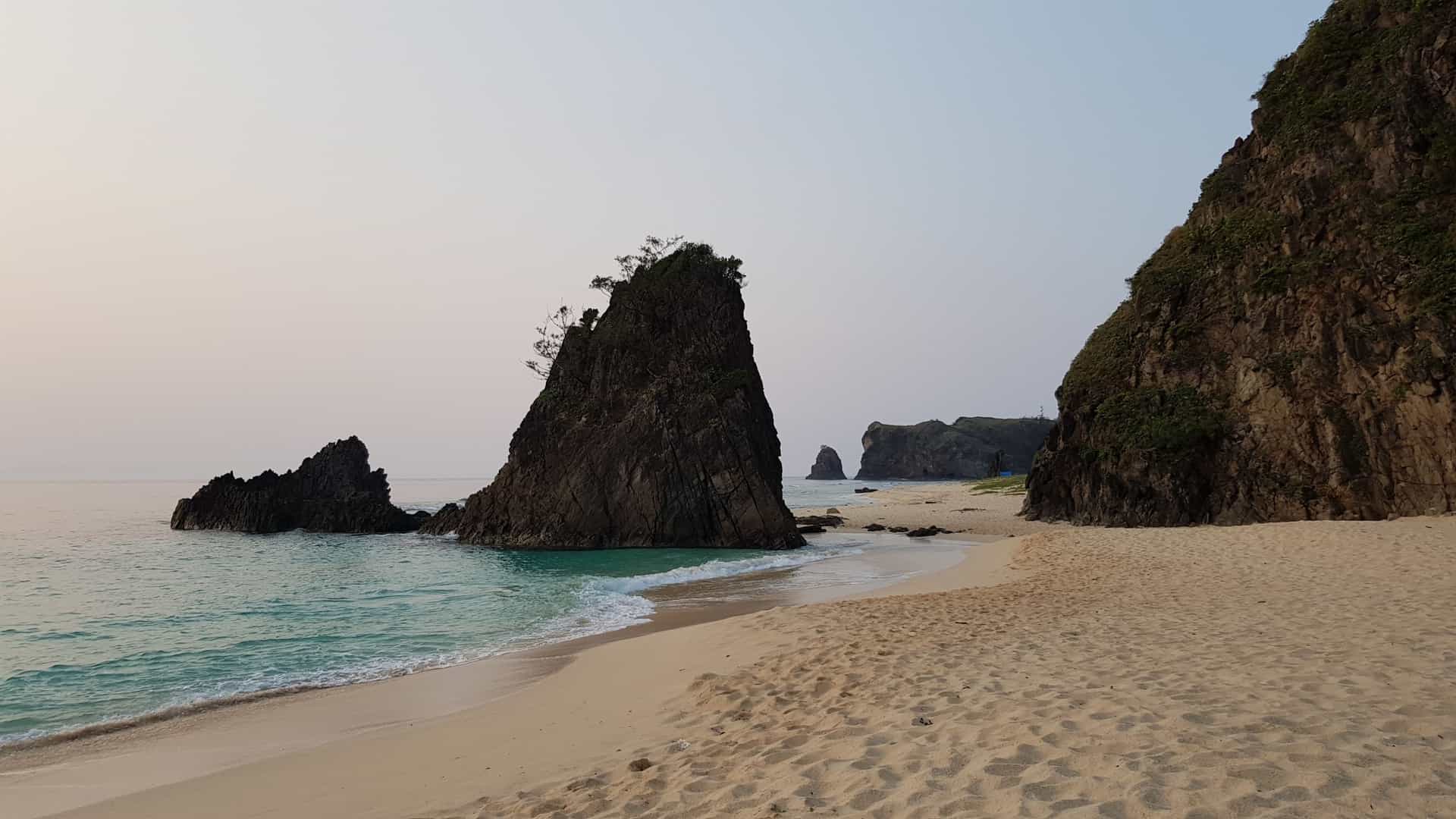 Large, dark, rocky sea stacks stand on the sandy beach and turquoise waters of Calayan Island, The Philippines.
