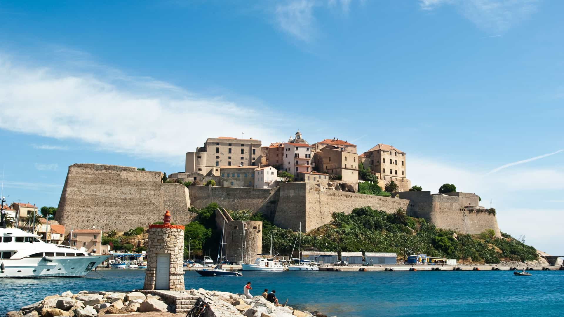 A stunning view of the historic Calvi Citadel on the Corsican coastline, with its fortified walls overlooking a vibrant harbor filled with boats and a clear blue sky.