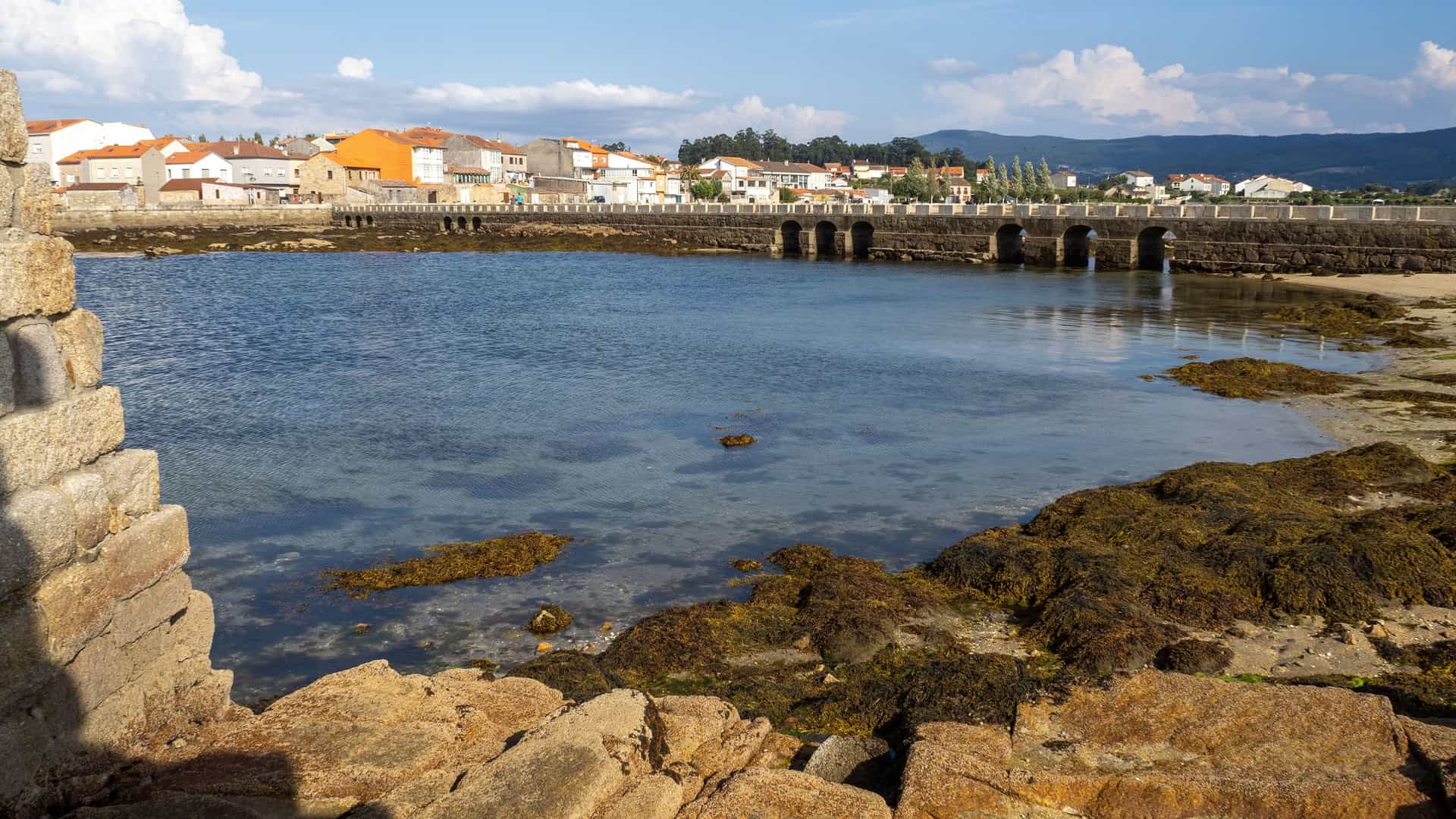 A view of the historic town of Cambados, Spain, across a shallow tidal inlet. A long stone bridge with multiple arches connects the town to the foreground, which is a rocky shoreline with patches of moss and seaweed. The town in the distance features white buildings with red-tiled roofs under a blue sky.
