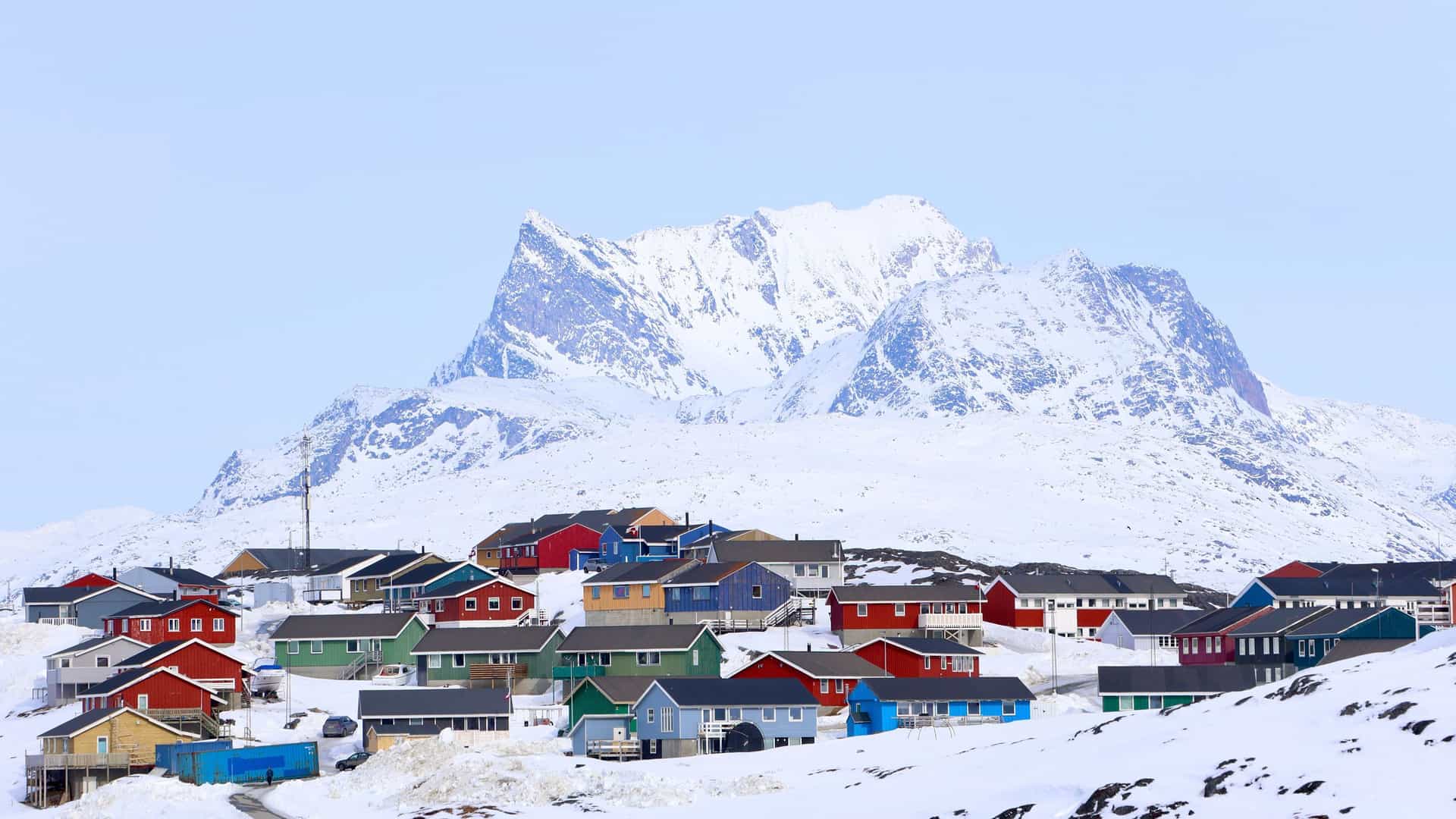A wide-angle landscape of the colorful houses of Camp Kangiusaq, Greenland, a small settlement surrounded by snow-covered terrain and towering, jagged mountains.