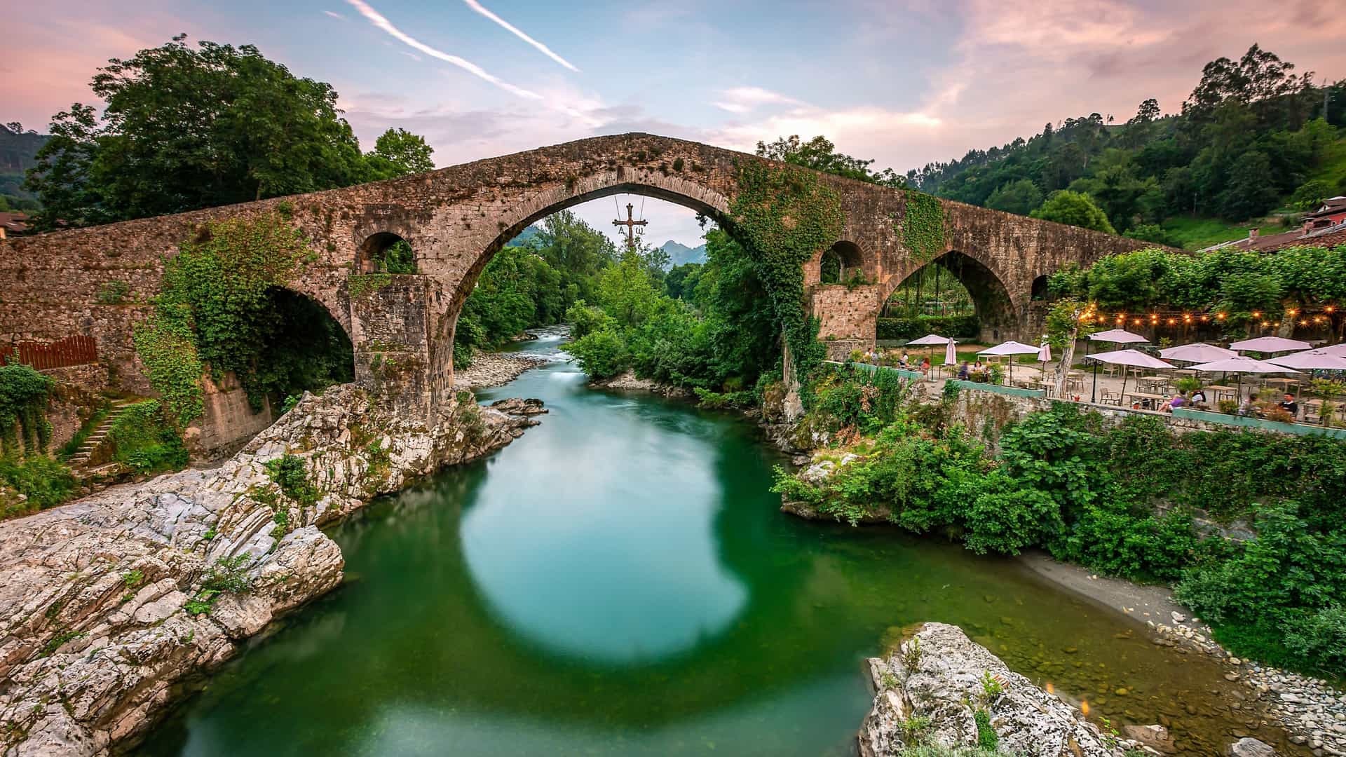 An ancient Roman bridge arches over a green river in Cangas de Onís, Spain, featuring a victory cross suspended in the center.