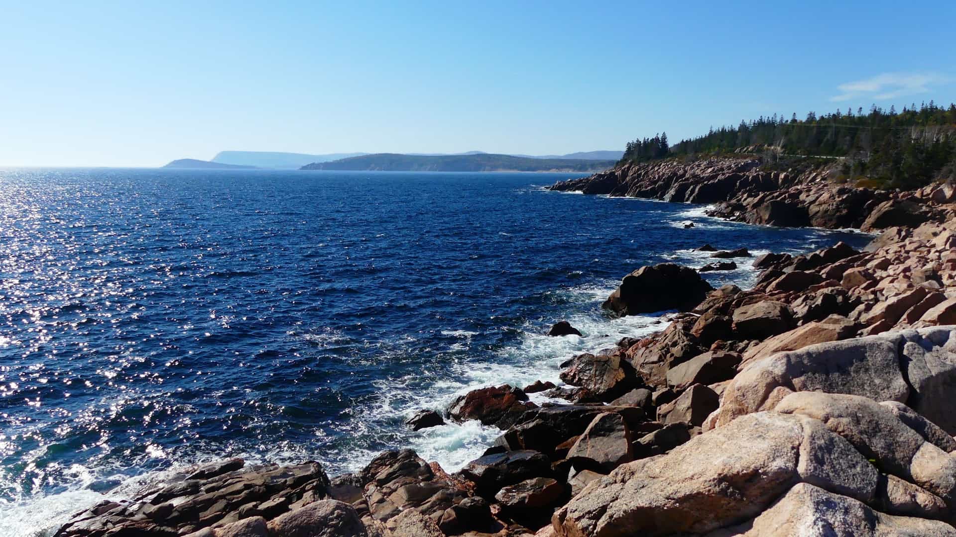 A dramatic coastal landscape on Cape Breton Island, Nova Scotia, Canada, shows a rocky shoreline meeting the sparkling deep blue Atlantic Ocean under a clear sky.