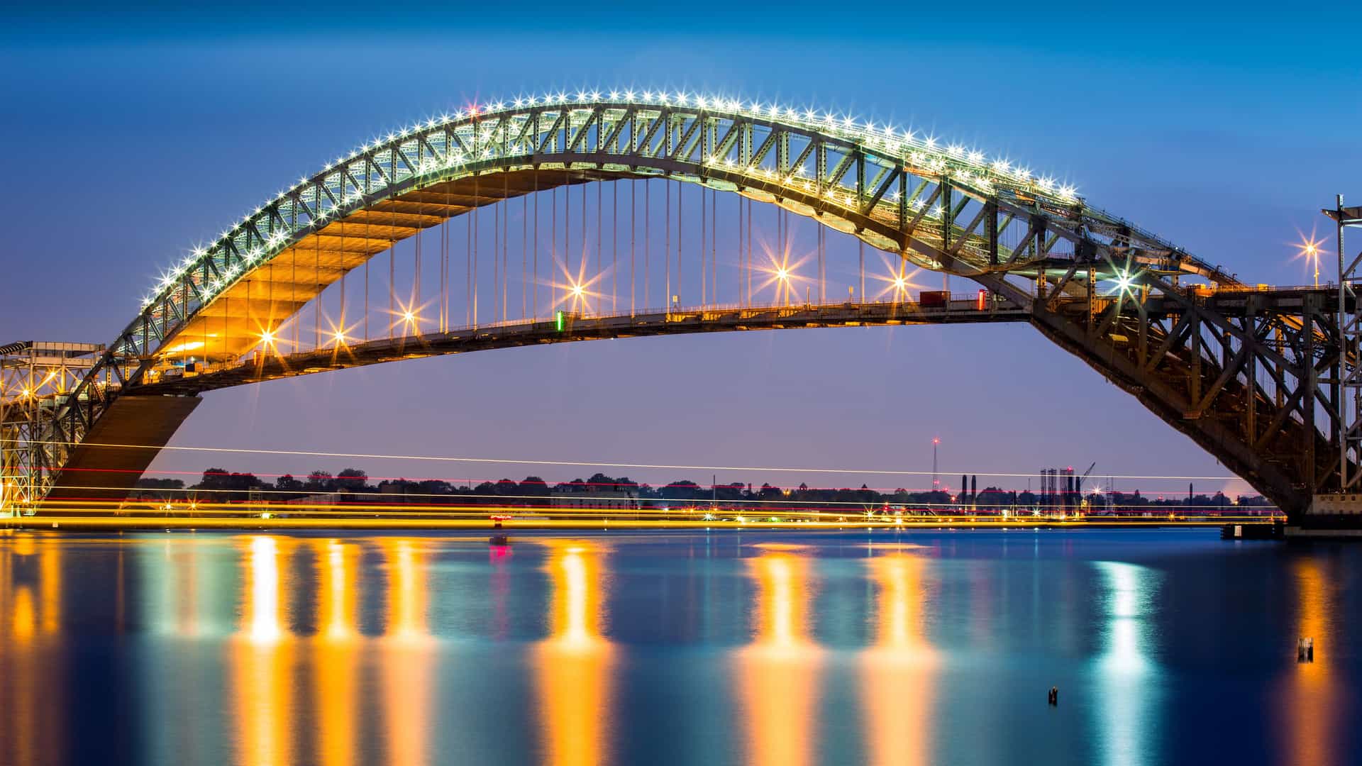 A night-time view of the iconic Bayonne Bridge in New Jersey, brightly lit against a dark sky, reflecting its lights on the still water of the Kill Van Kull waterway.