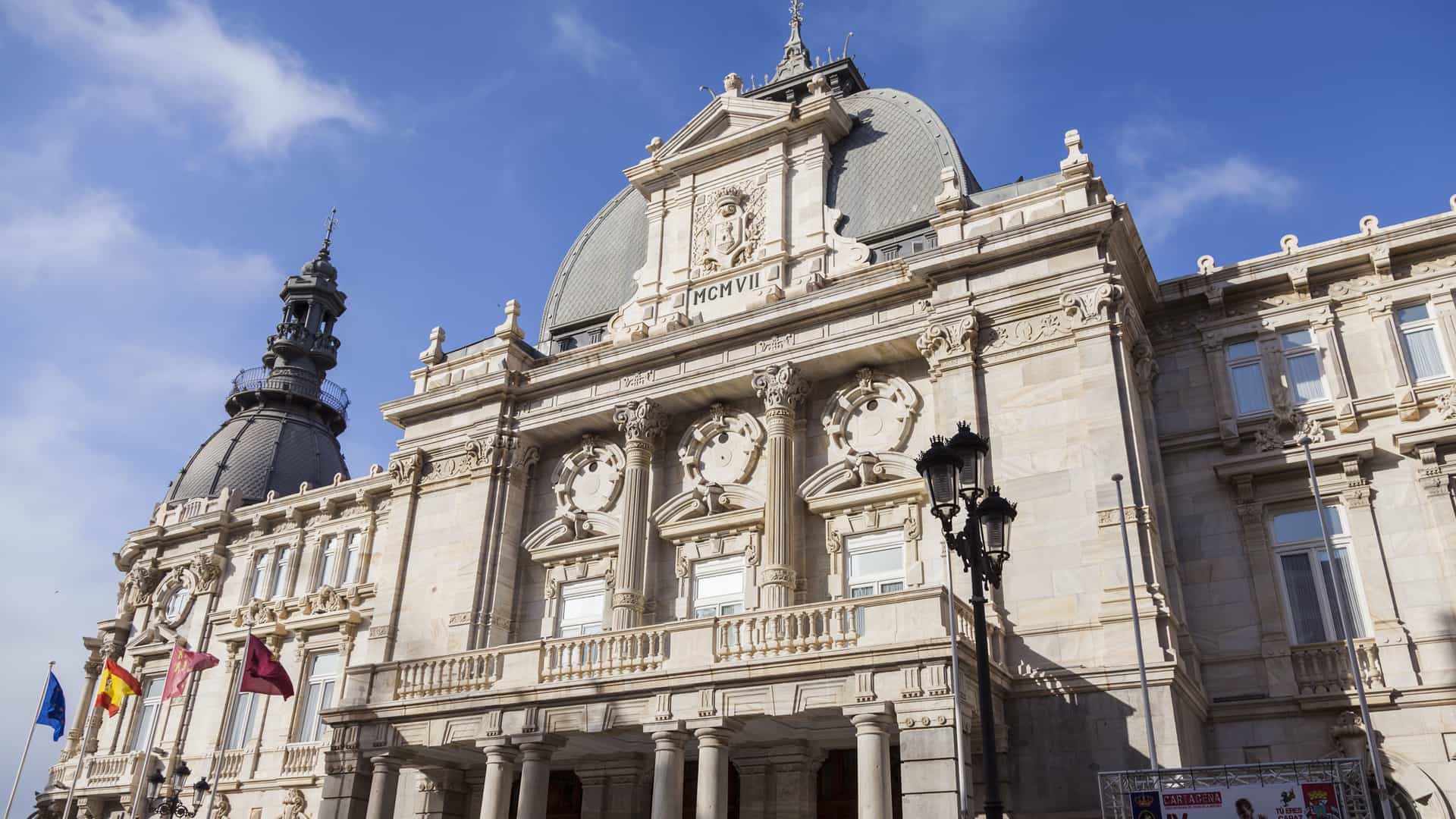 A low-angle shot of the magnificent Palacio Consistorial, the town hall of Cartagena, Spain, showcasing its ornate, white marble facade and domed rooftops against a blue sky.