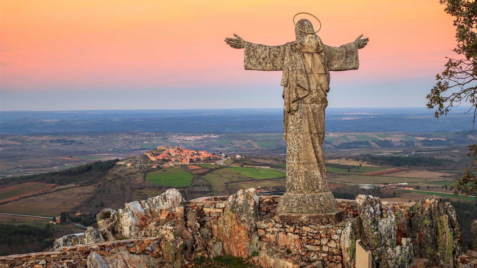 A stone statue of Jesus Christ with outstretched arms overlooks the small village of Castelo Rodrigo, Portugal, with a stunning sunset in the background.