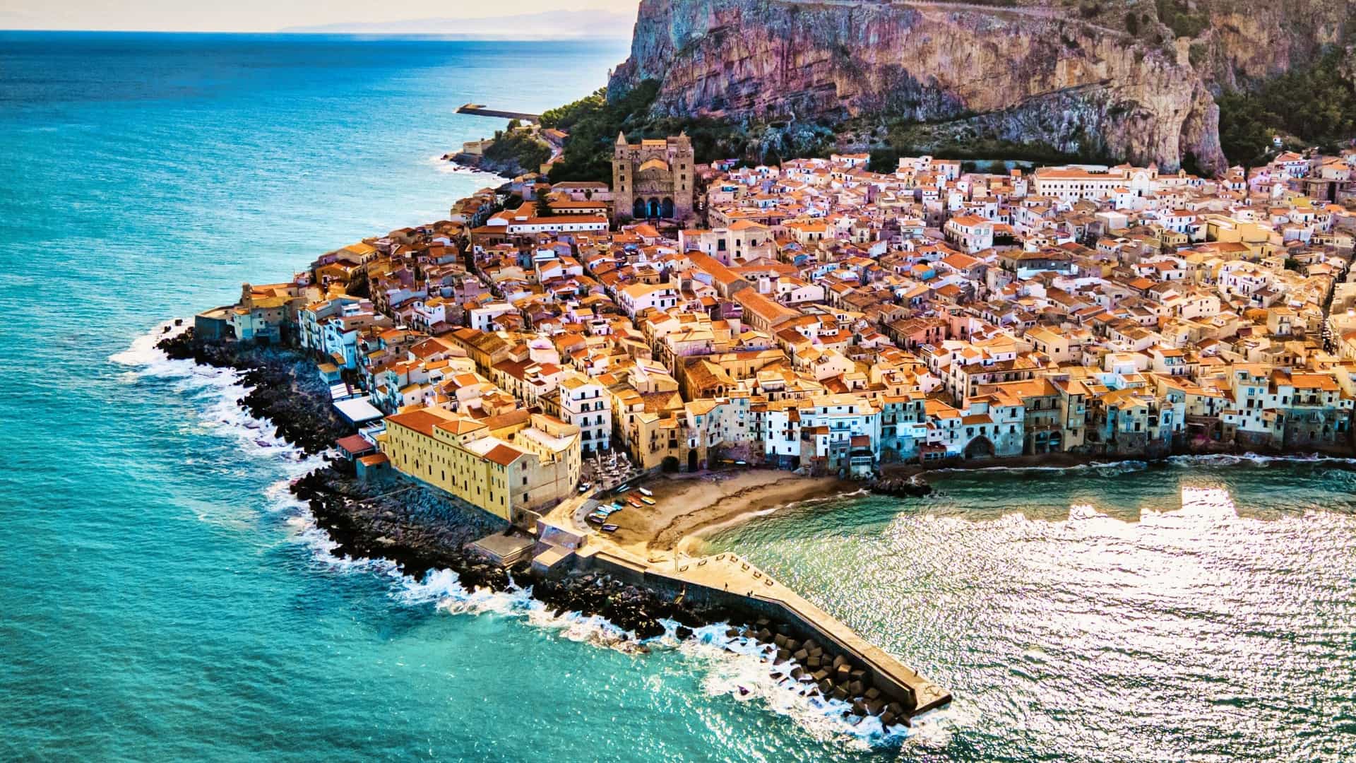 An aerial view of Cefalù, Sicily, Italy, shows the densely packed old town with orange-tiled roofs nestled against the turquoise Mediterranean Sea and the massive cliff of Rocca di Cefalù.