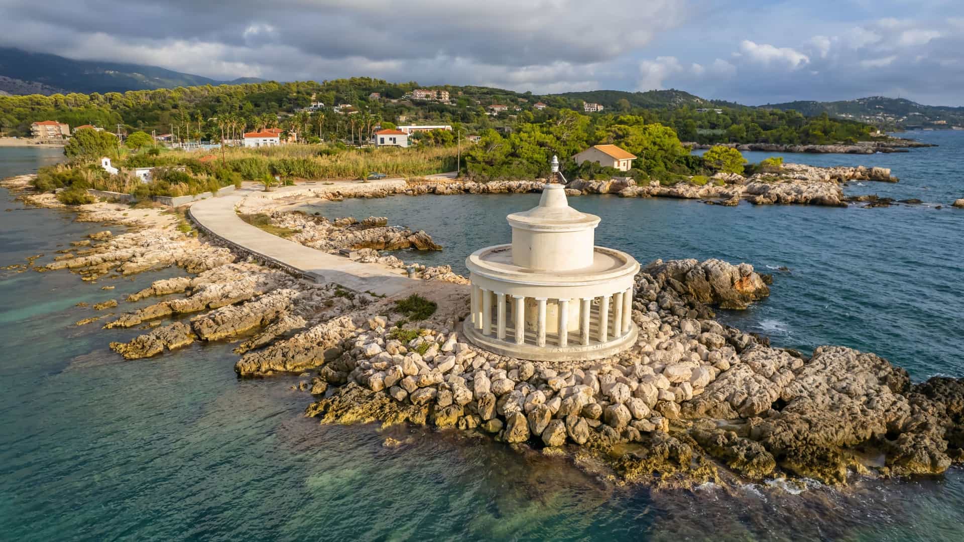 An aerial shot of the historic Fanari Lighthouse on a small rocky islet, connected by a causeway to the mainland of Argostoli, Cephalonia, with villas and lush hills in the background.