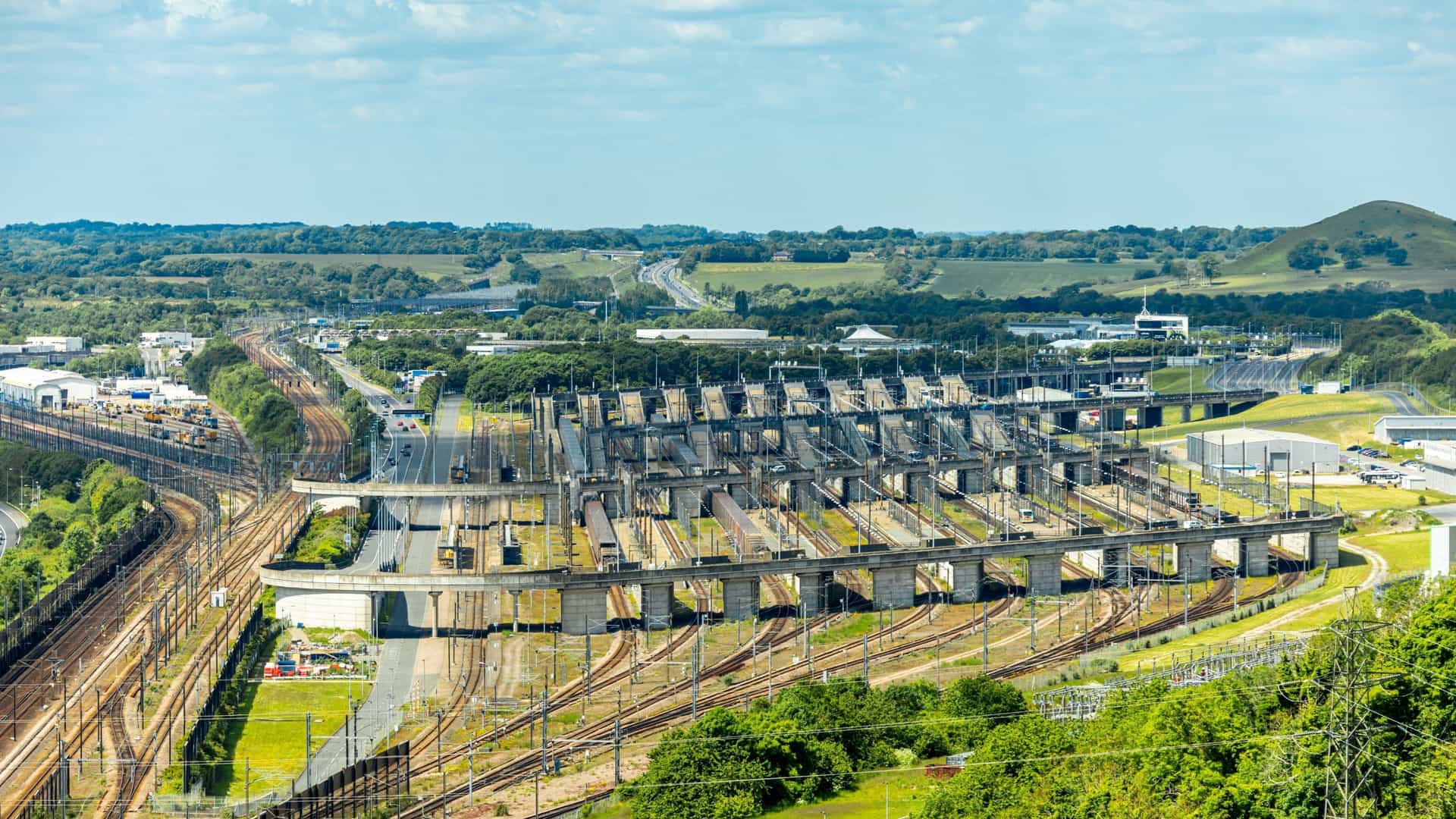 An aerial view of the Channel Tunnel's Folkestone terminal in the UK, showcasing an intricate network of rail lines, loading platforms, and surrounding green hills.