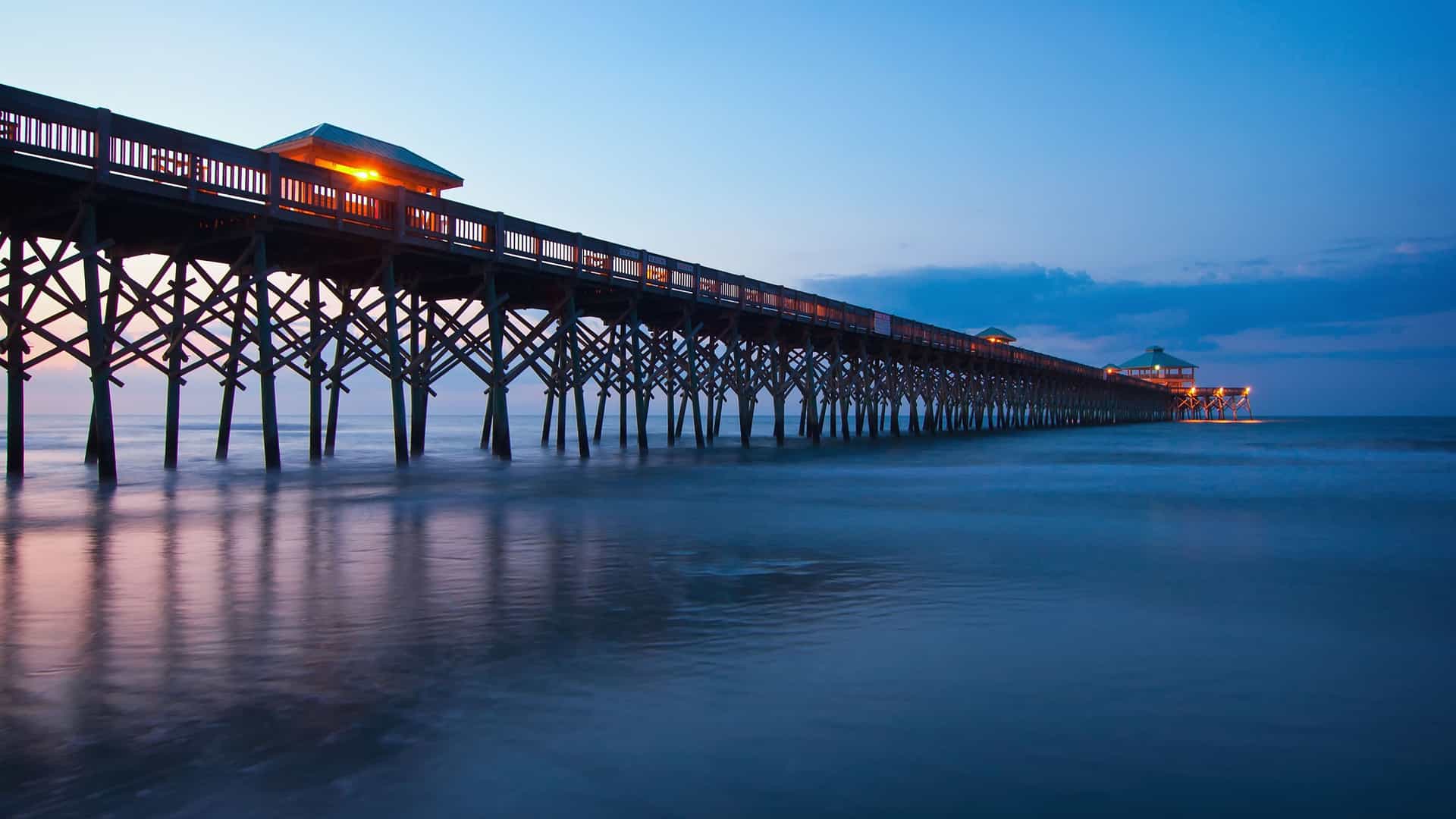 A stunning long exposure photograph of the Folly Beach Pier in Charleston, South Carolina, illuminated at dusk, with the calm ocean water and deep blue sky.