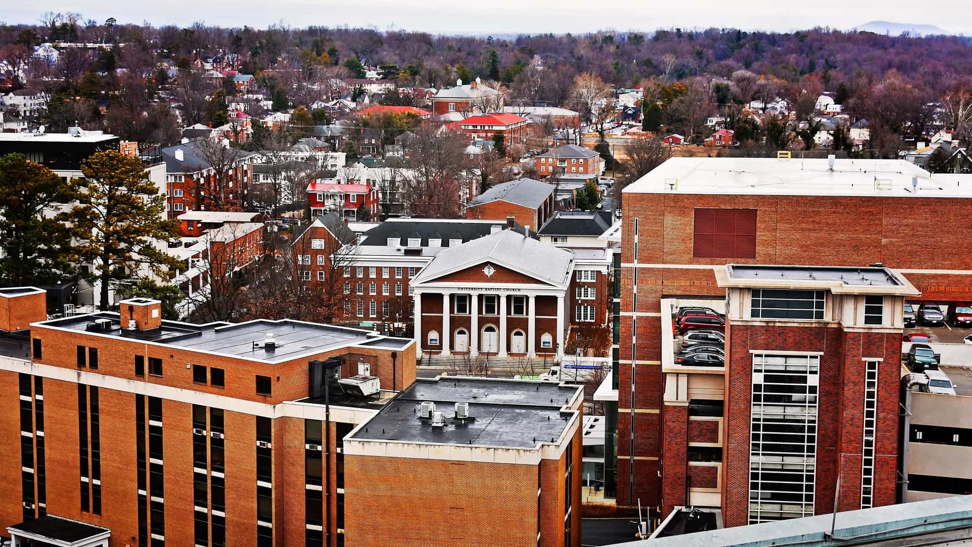 An elevated view of Charlottesville, Virginia, shows a mix of brick academic and residential buildings, with a prominent white building featuring columns in the center.