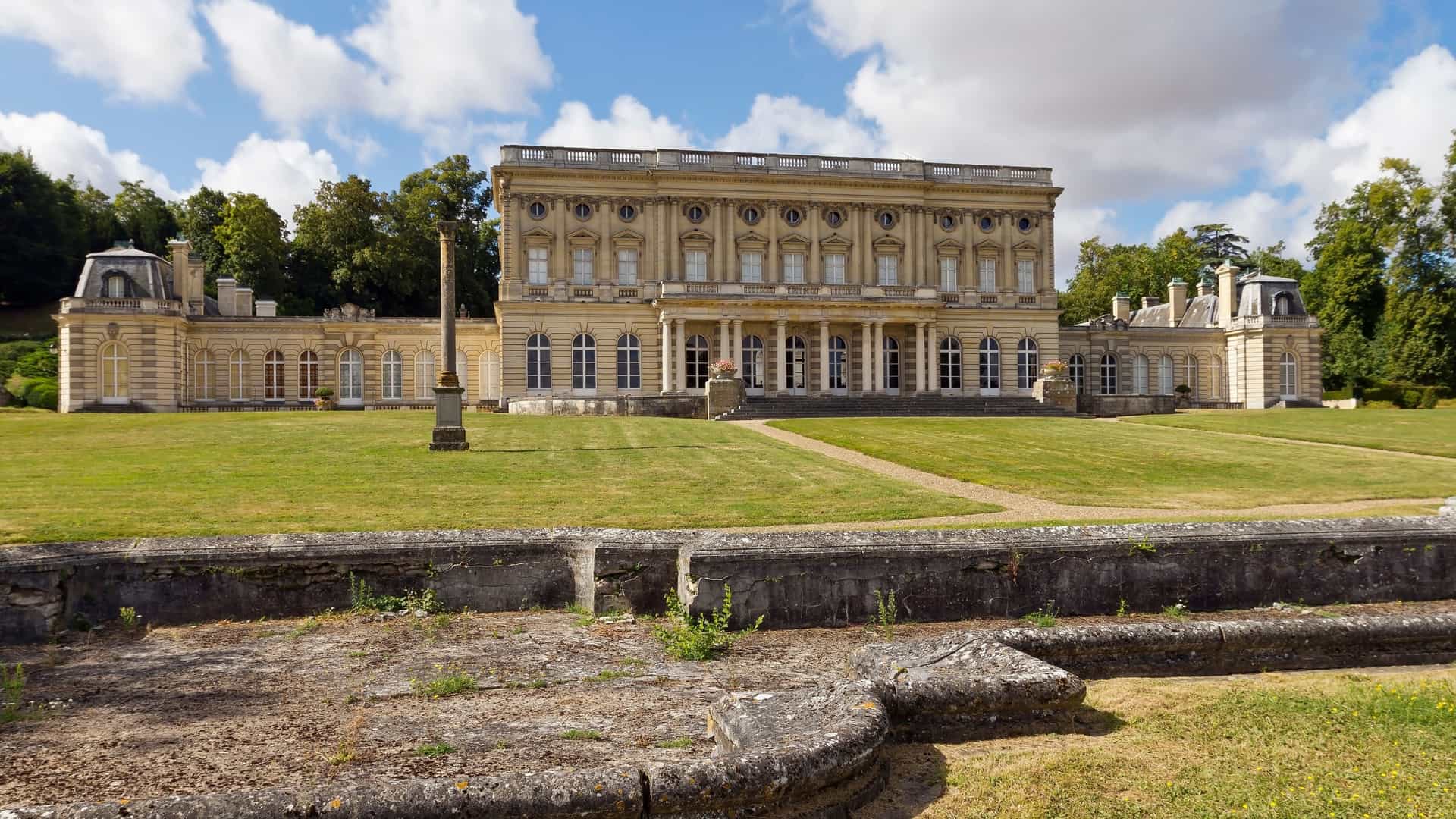 A grand, horizontal neoclassical building, the Château de Bizy in Vernon, France, is framed by expansive green lawns and a partly cloudy blue sky.