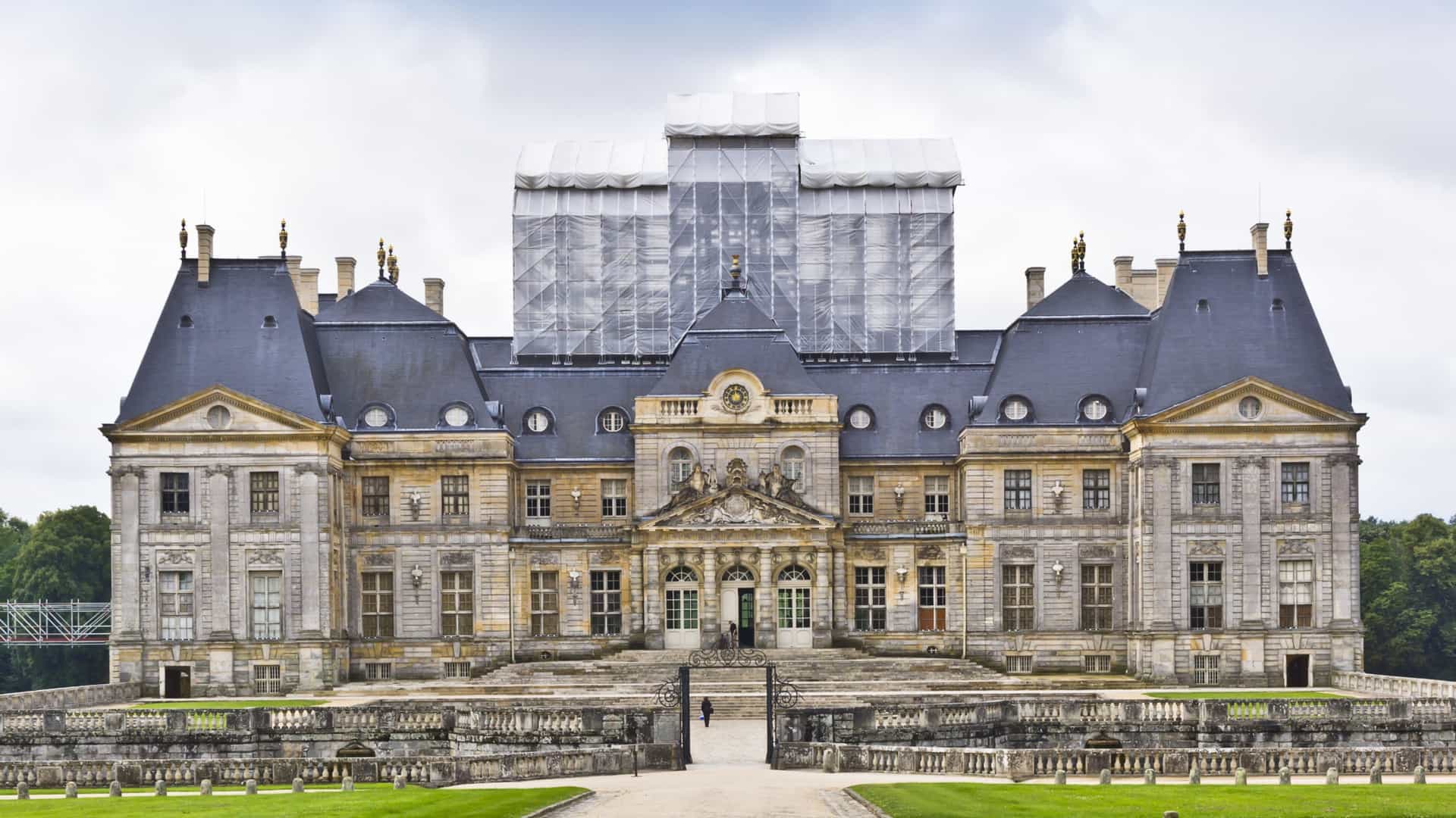 A view of the immense, symmetrical Baroque Château de Vaux-le-Vicomte in Maincy, France, with its prominent blue slate roofs and dormer windows. The central part of the château is covered in scaffolding and white construction material, set against a green lawn and a cloudy sky.