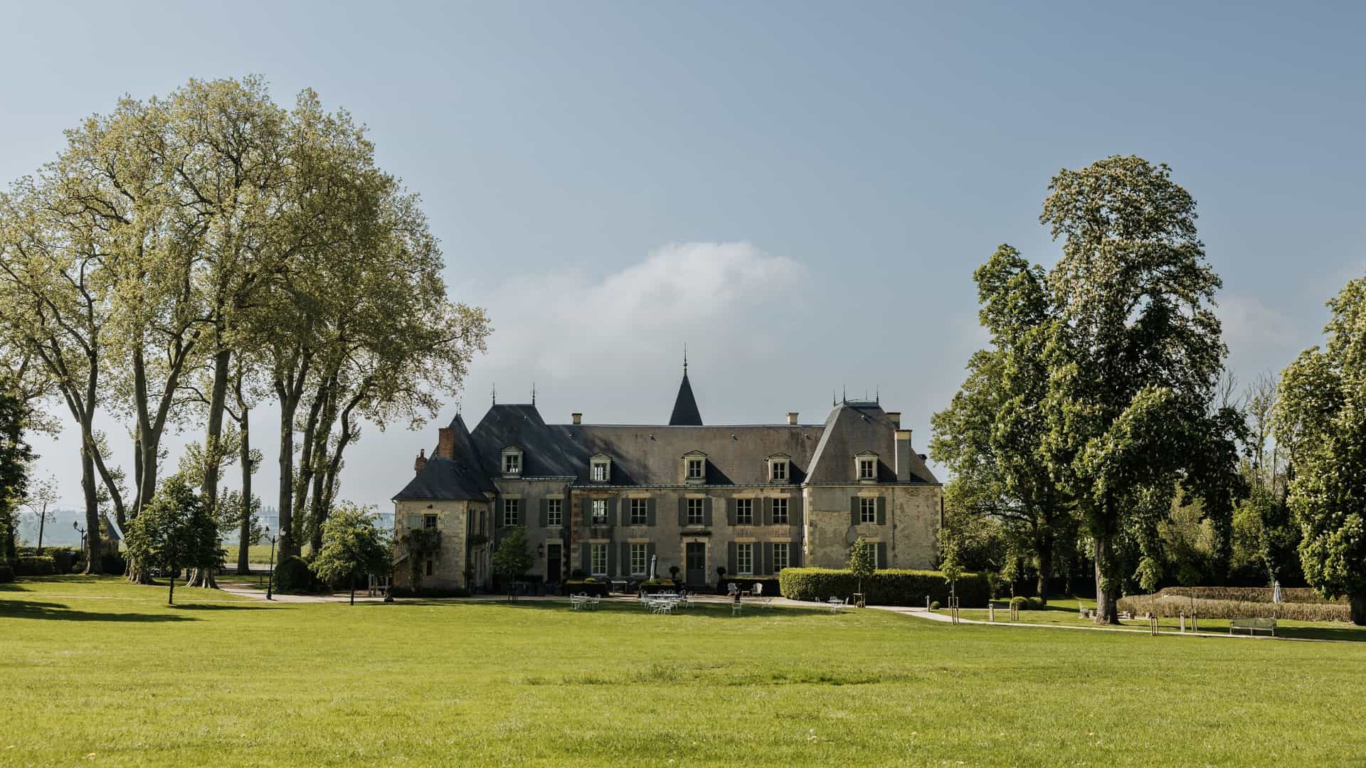 A picturesque view of the Château du Taillis, France, shows a stately, traditional stone château with a steep slate roof set back on a vast, sunny green lawn, framed by tall, mature trees.