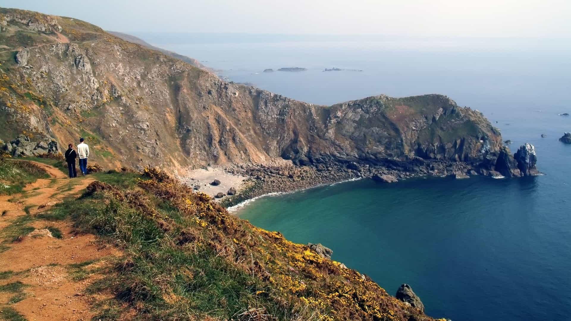 A high-angle view of a couple standing on a winding cliff path overlooking a rocky coastline and the sea near Cherbourg, France.